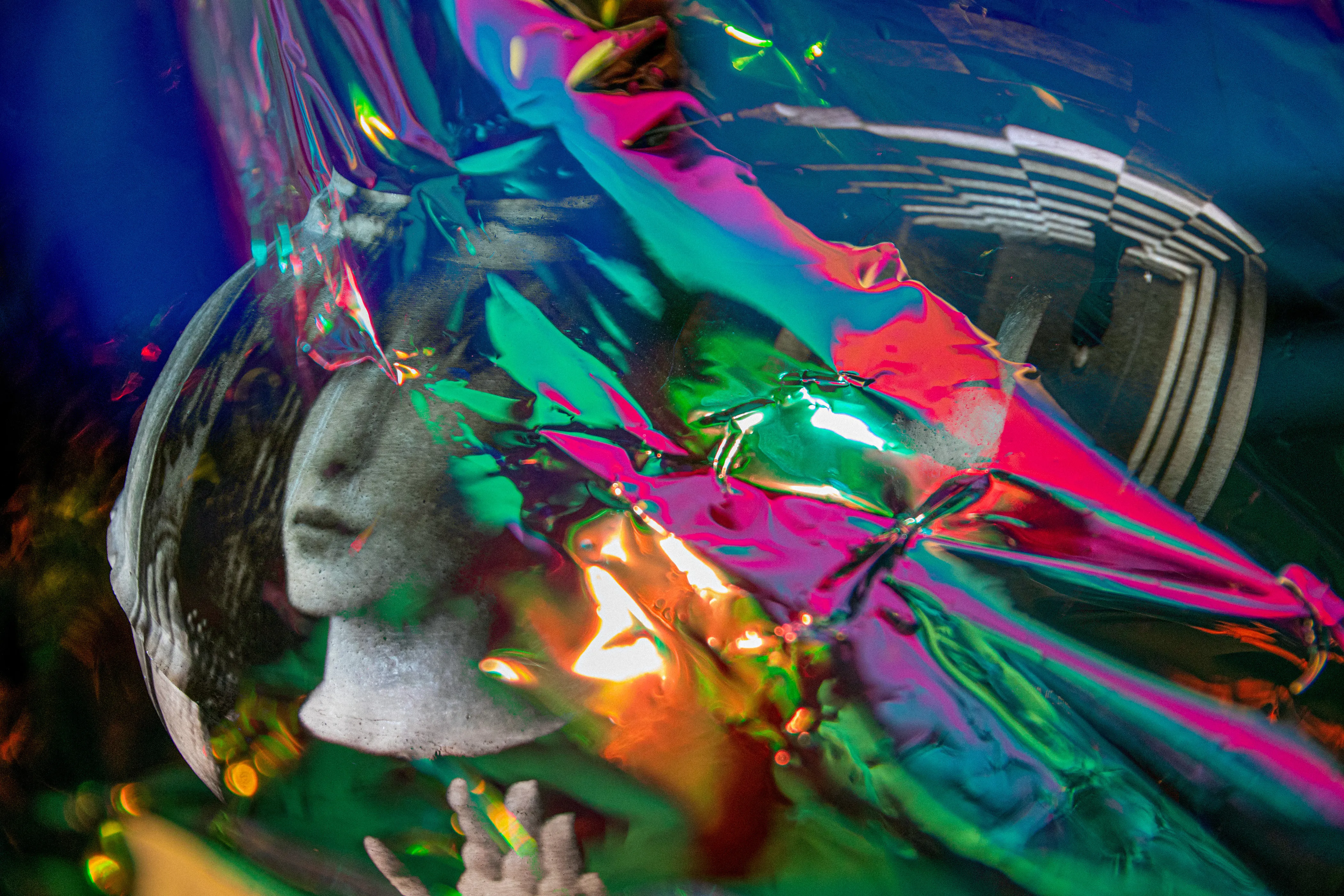 A pale sculpted head turned to the left inside a clear round bowl, with a hand at the bottom, among crinkled pink, green and blue foil and a ribbed pattern at the upper right.