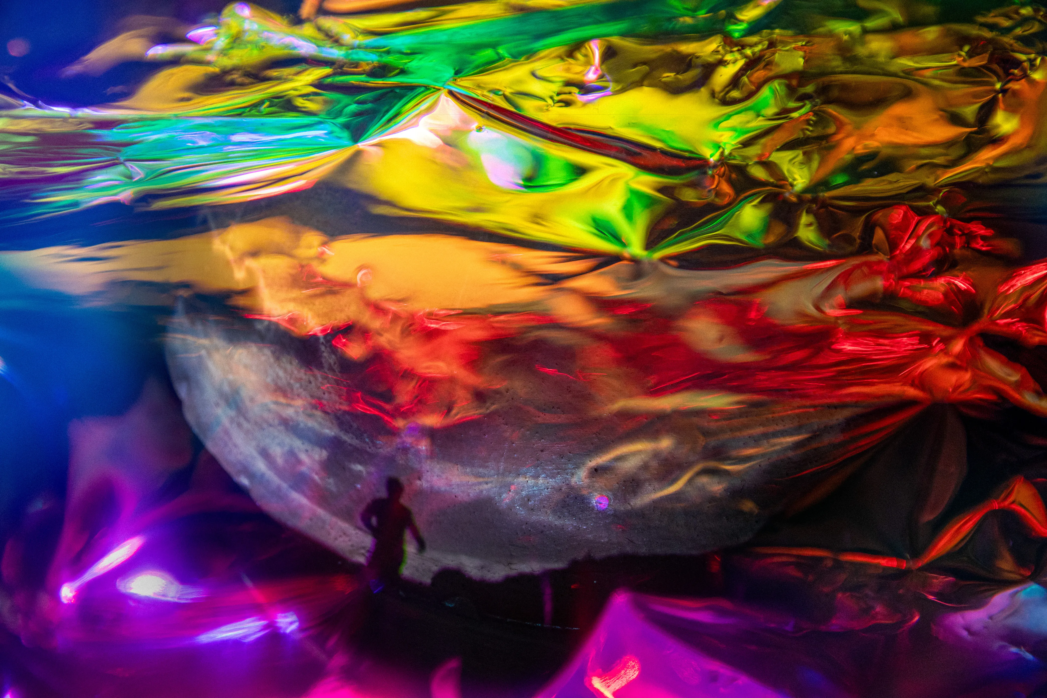Folded metallic foil in orange, yellow, green and purple over a pale rounded form, with a small dark standing figure at the lower center.