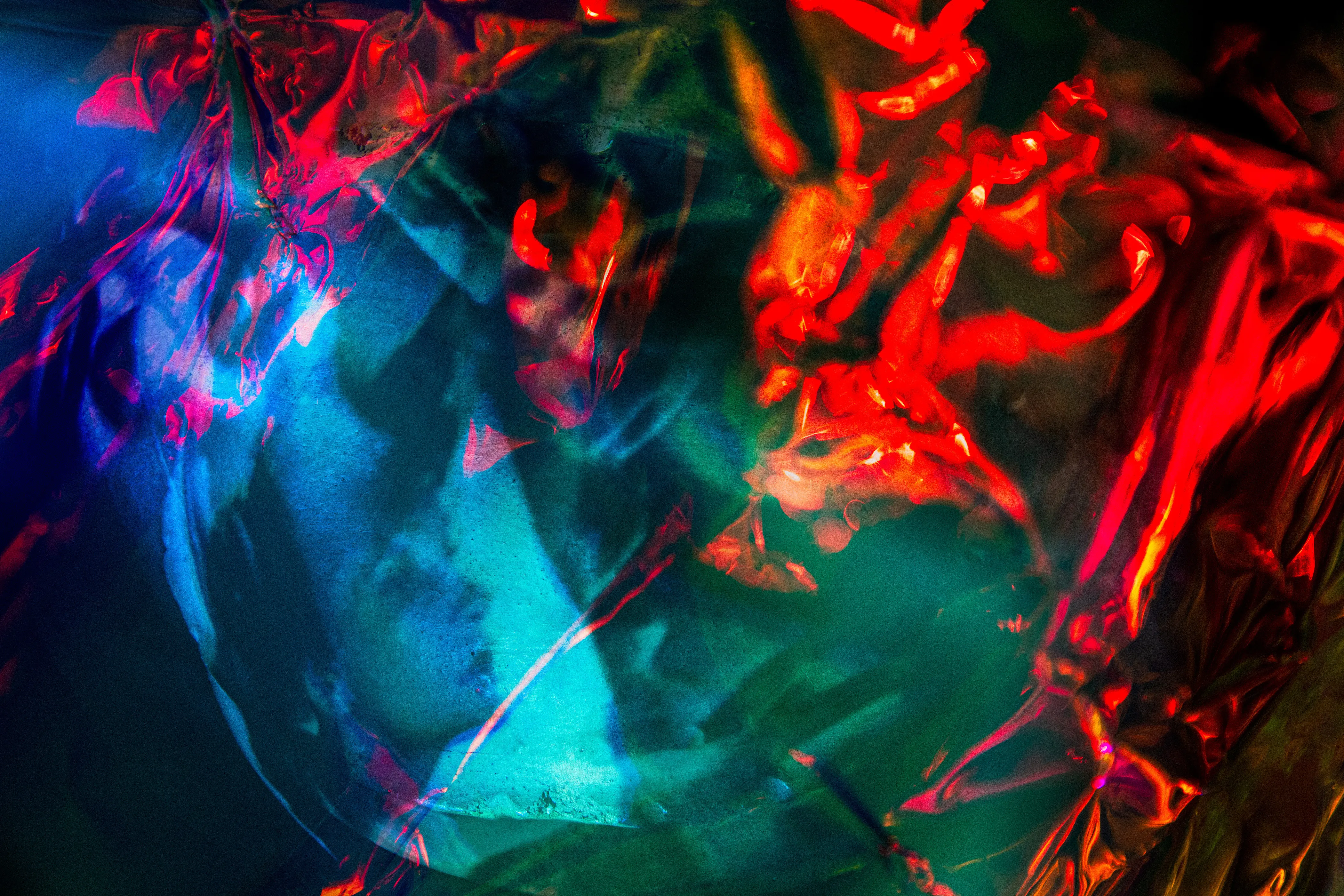 Red crumpled foil at the right and a blue and turquoise form at the left, with a pale face barely visible at center left.