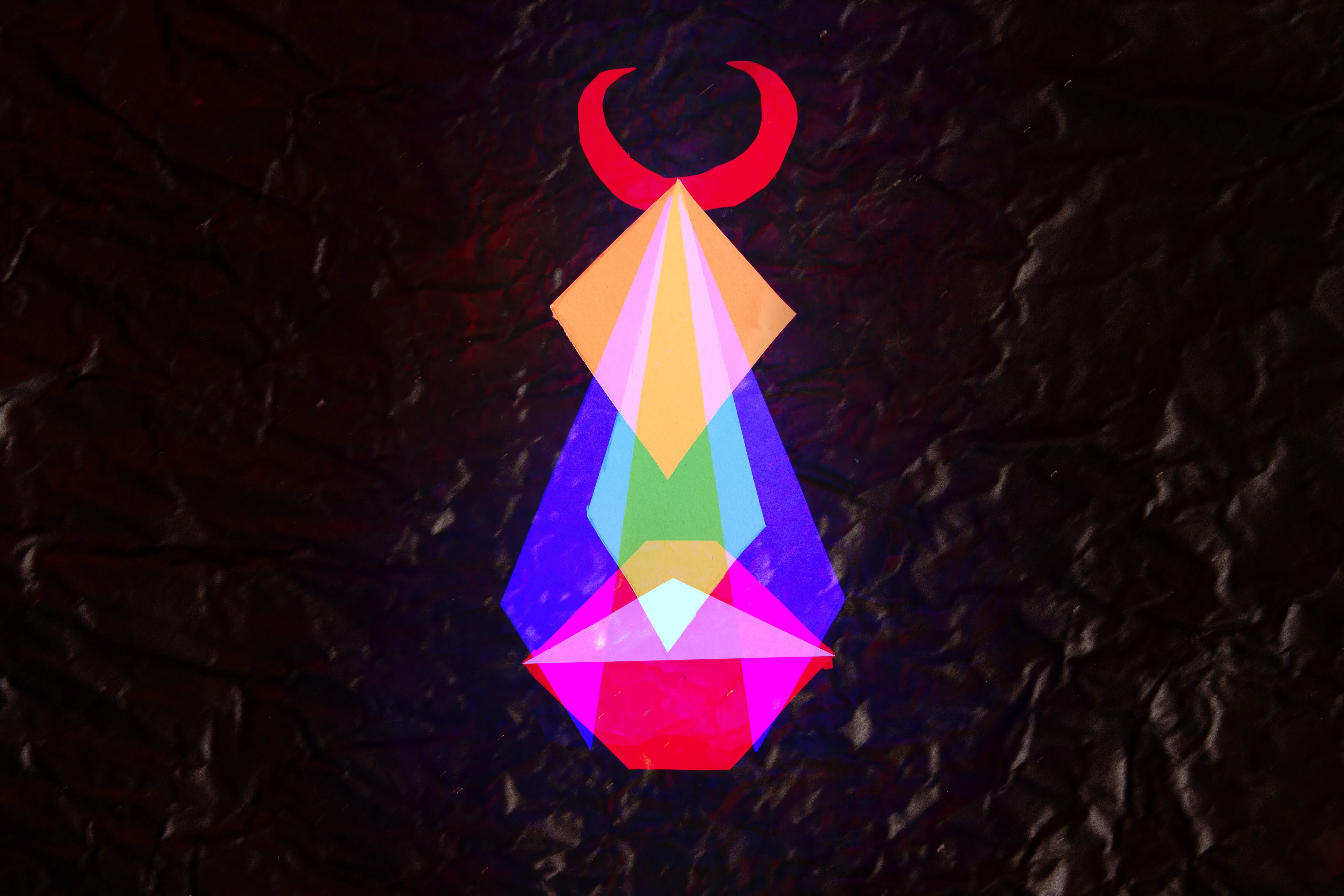 A symmetrical stack of translucent diamond and triangle shapes in blue, magenta, yellow and green, topped by a red crescent, on a dark crinkled ground.