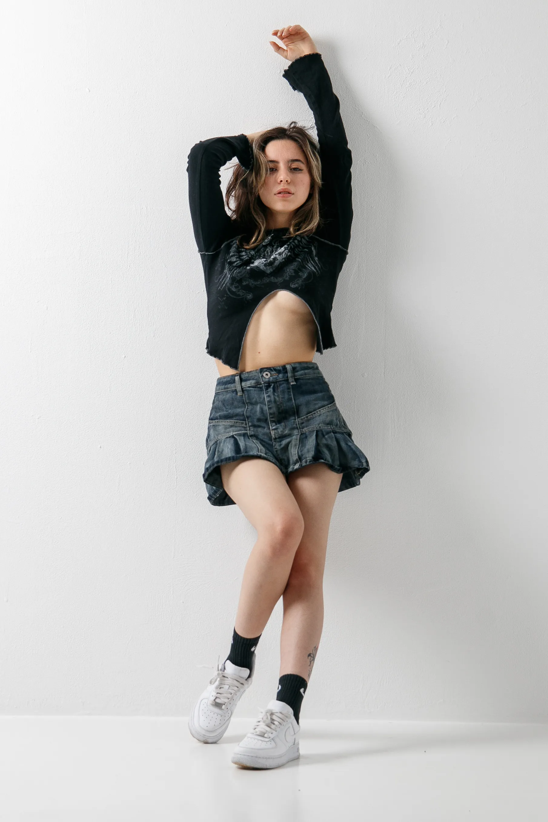 A woman leans against a white wall with one arm raised overhead, in a black cropped long-sleeved top, a denim ruffled mini skirt and white sneakers.