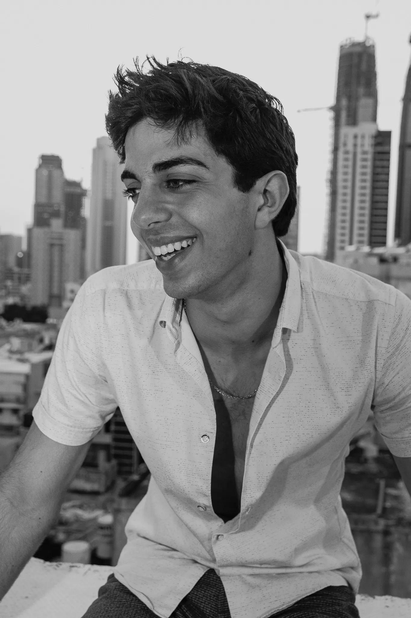 A black and white portrait of a smiling young man in an open short-sleeved shirt, looking to the left, with high-rise buildings behind him.