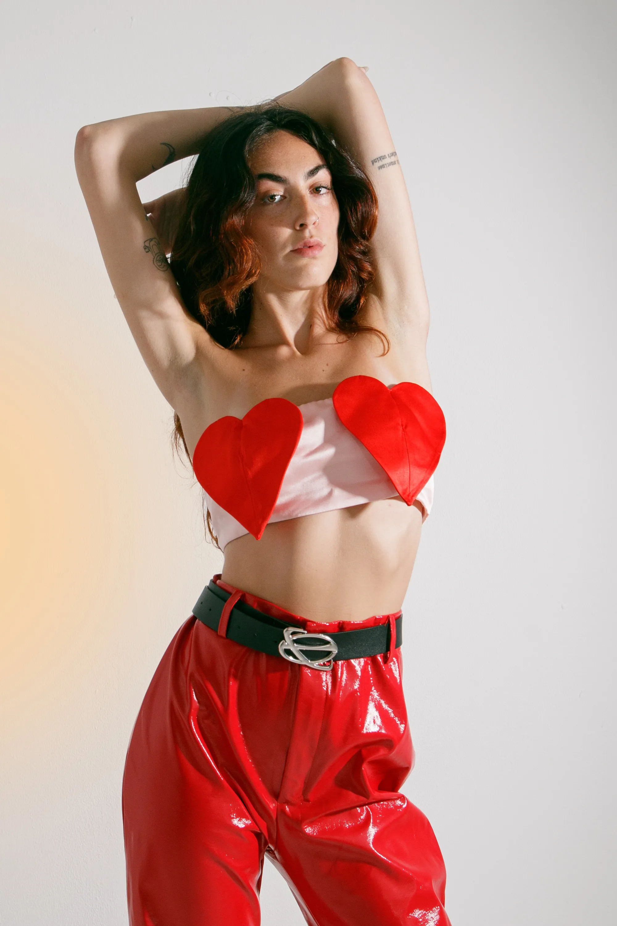 A closer view of the same outfit, with the woman's arms raised over her head, red heart-shaped pieces on her chest and glossy red trousers.