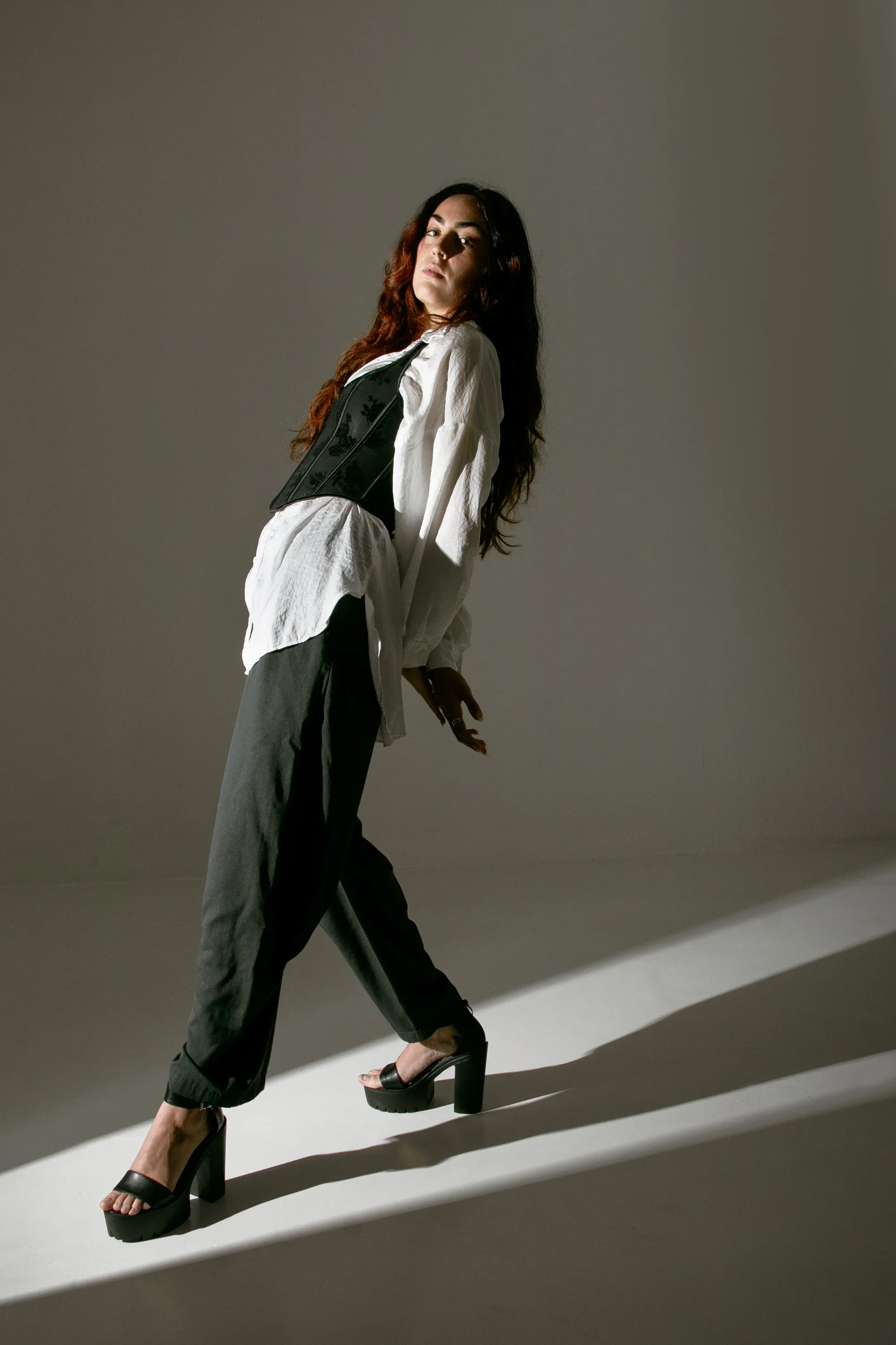A woman in a white shirt, dark vest and trousers and platform sandals leans back at an angle, her long hair falling behind, on a gray studio floor crossed by a band of light.