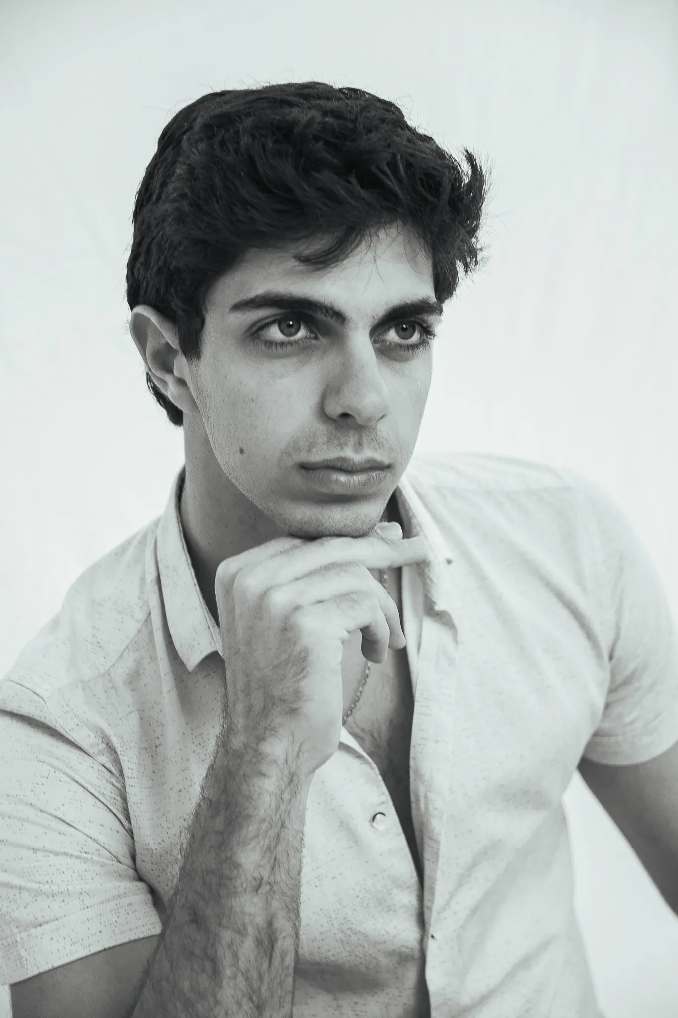 A black and white portrait of a young man with dark hair resting his chin on his hand, in a short-sleeved shirt.