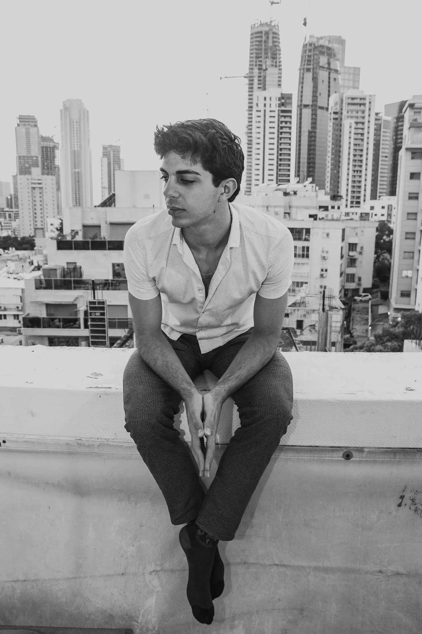 A black and white photograph of a young man sitting on a rooftop ledge, hands clasped between his knees, with city towers behind him.