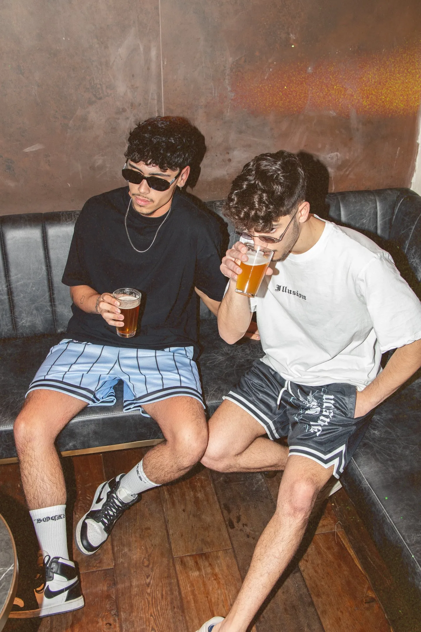 Two young men sit on a black leather bench, one in sunglasses and a black T-shirt, the other in a white T-shirt, both drinking from glasses of beer.