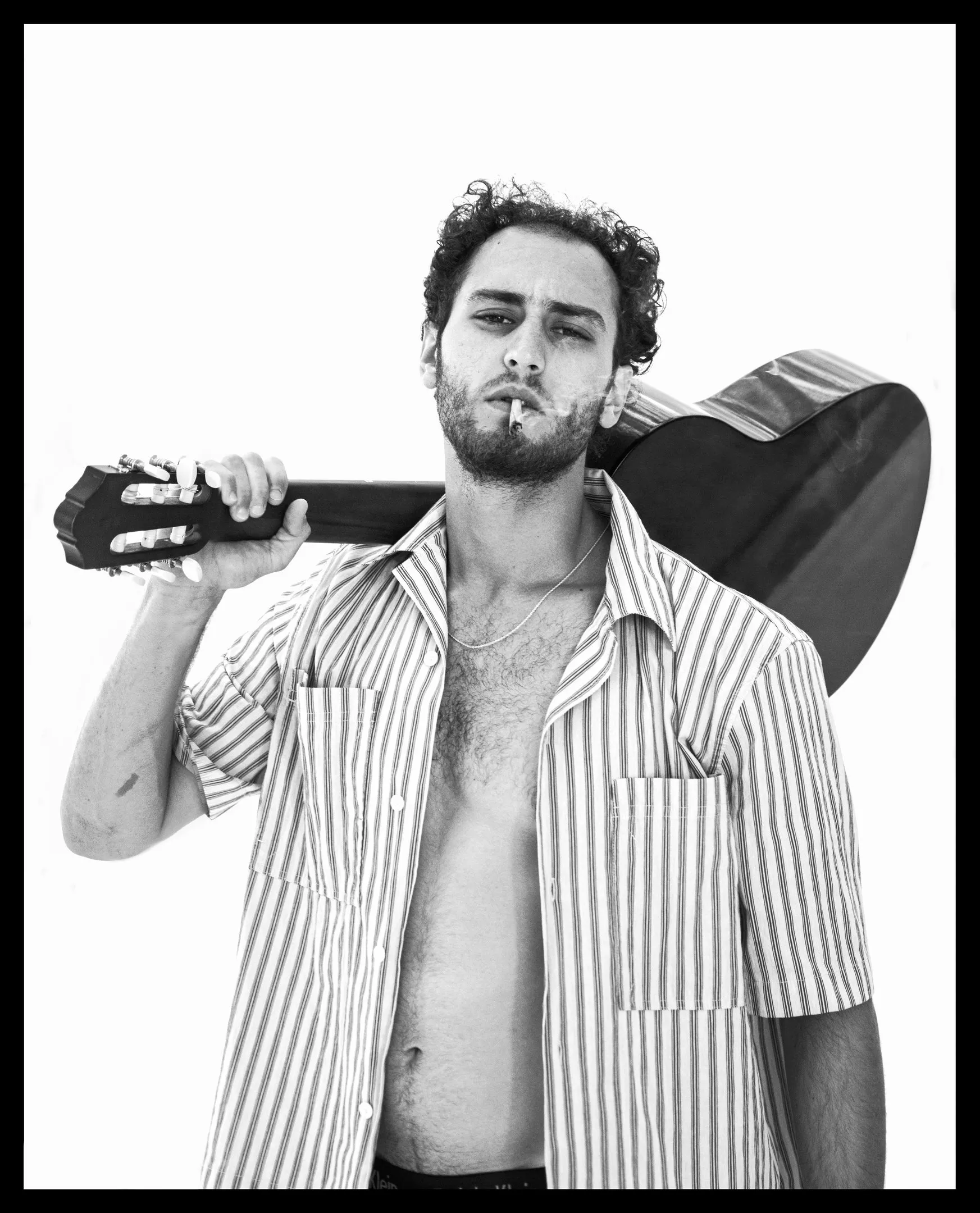 A man with curly dark hair and a beard, a cigarette in his mouth, holds an acoustic guitar over his shoulder, in an open striped shirt, against white.