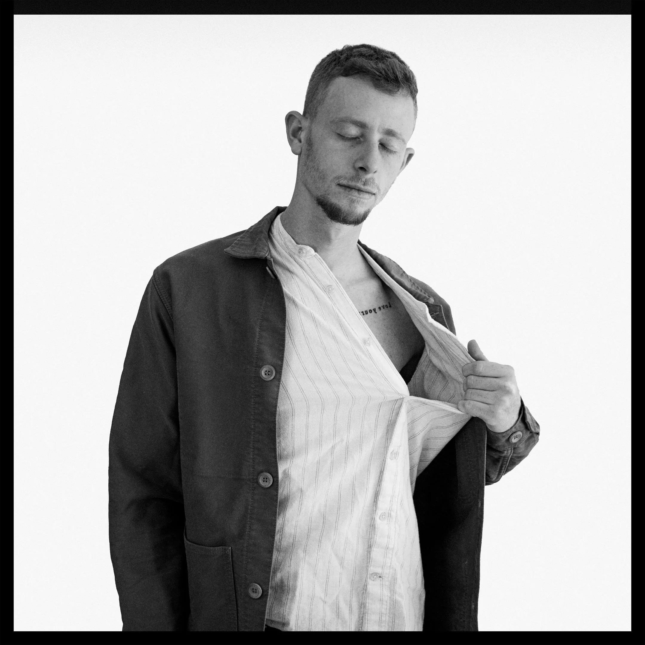 A young man looking down pulls open a striped shirt under a dark jacket, a small tattoo showing at his chest.