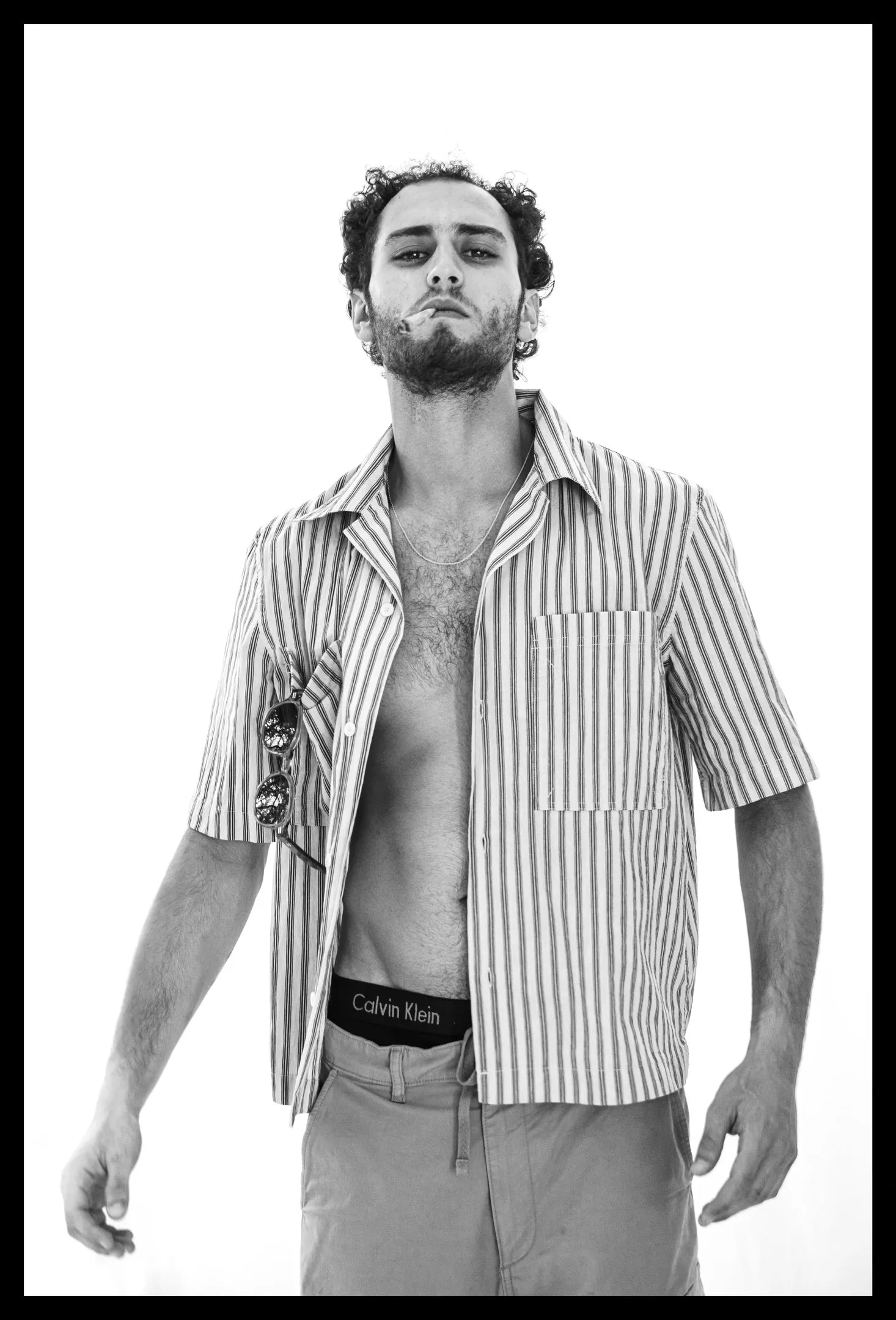 A man with curly hair and a beard stands with arms at his sides in an open striped short-sleeved shirt and light trousers, the waistband of his underwear showing.