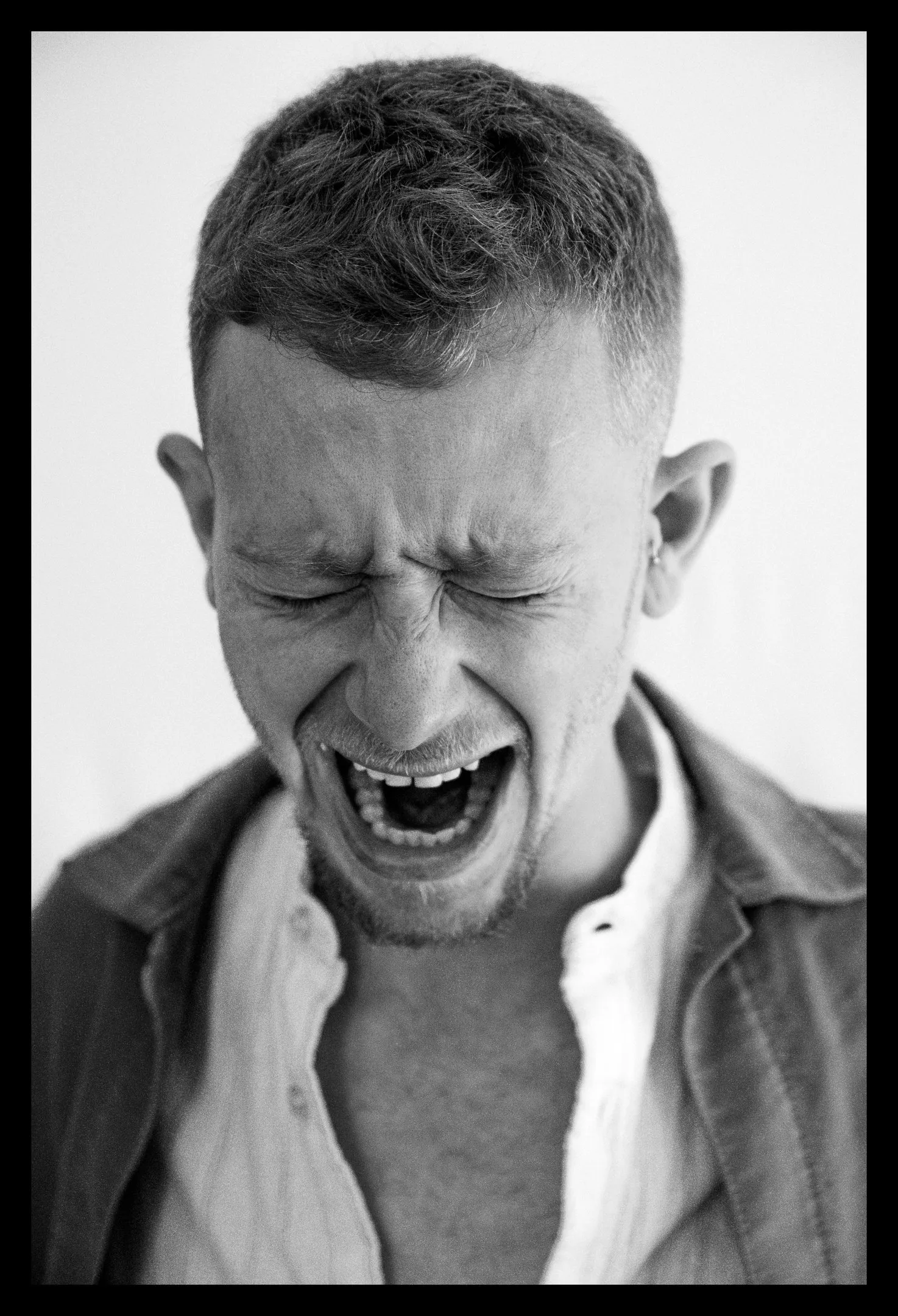 A close portrait of a man with short hair, eyes shut and mouth wide open, in an open dark jacket.