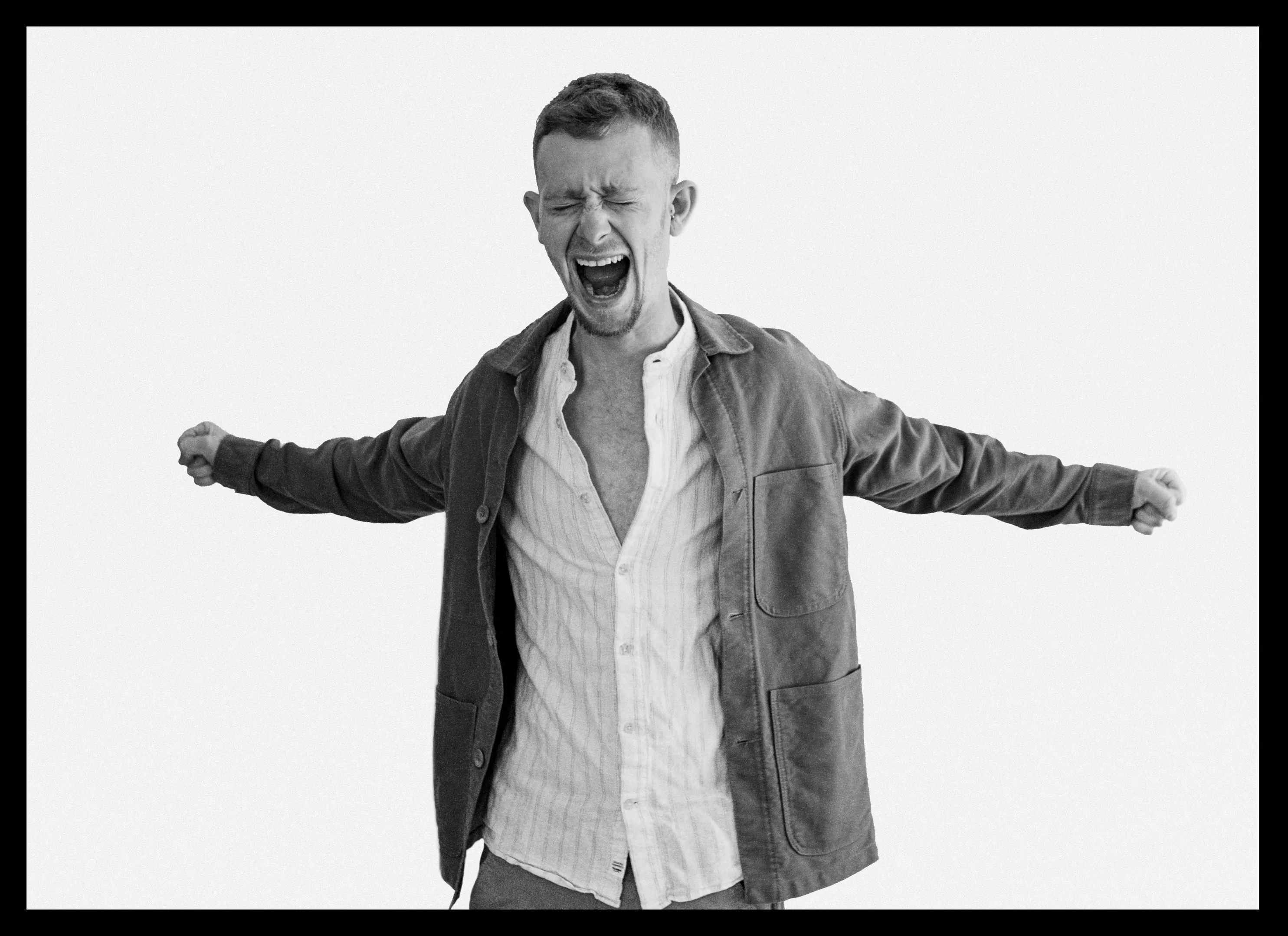 A man in an open dark jacket stands with arms stretched out to the sides and mouth open.