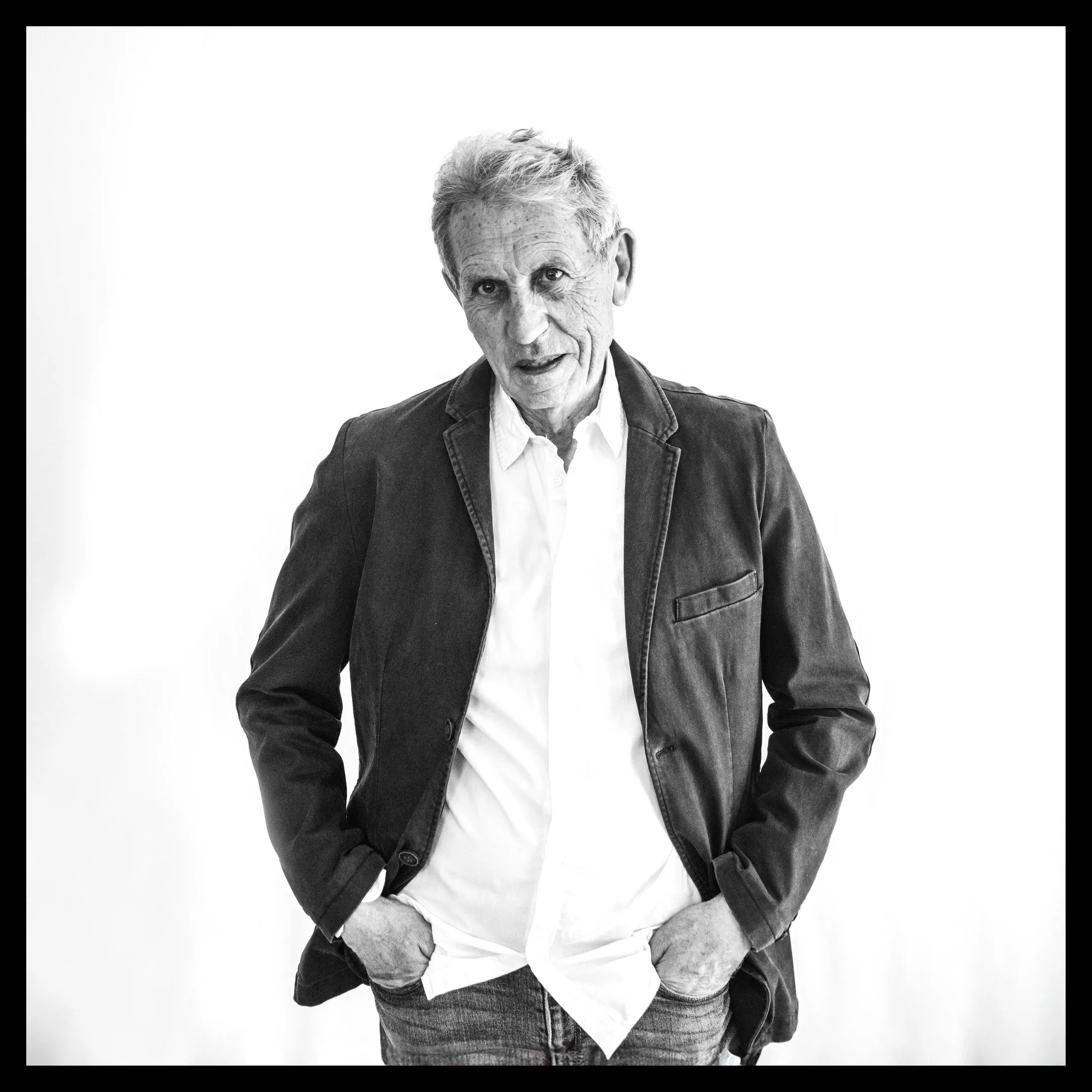 An older man with gray hair stands with his hands in his jeans pockets, in a dark jacket and white shirt, looking at the camera.