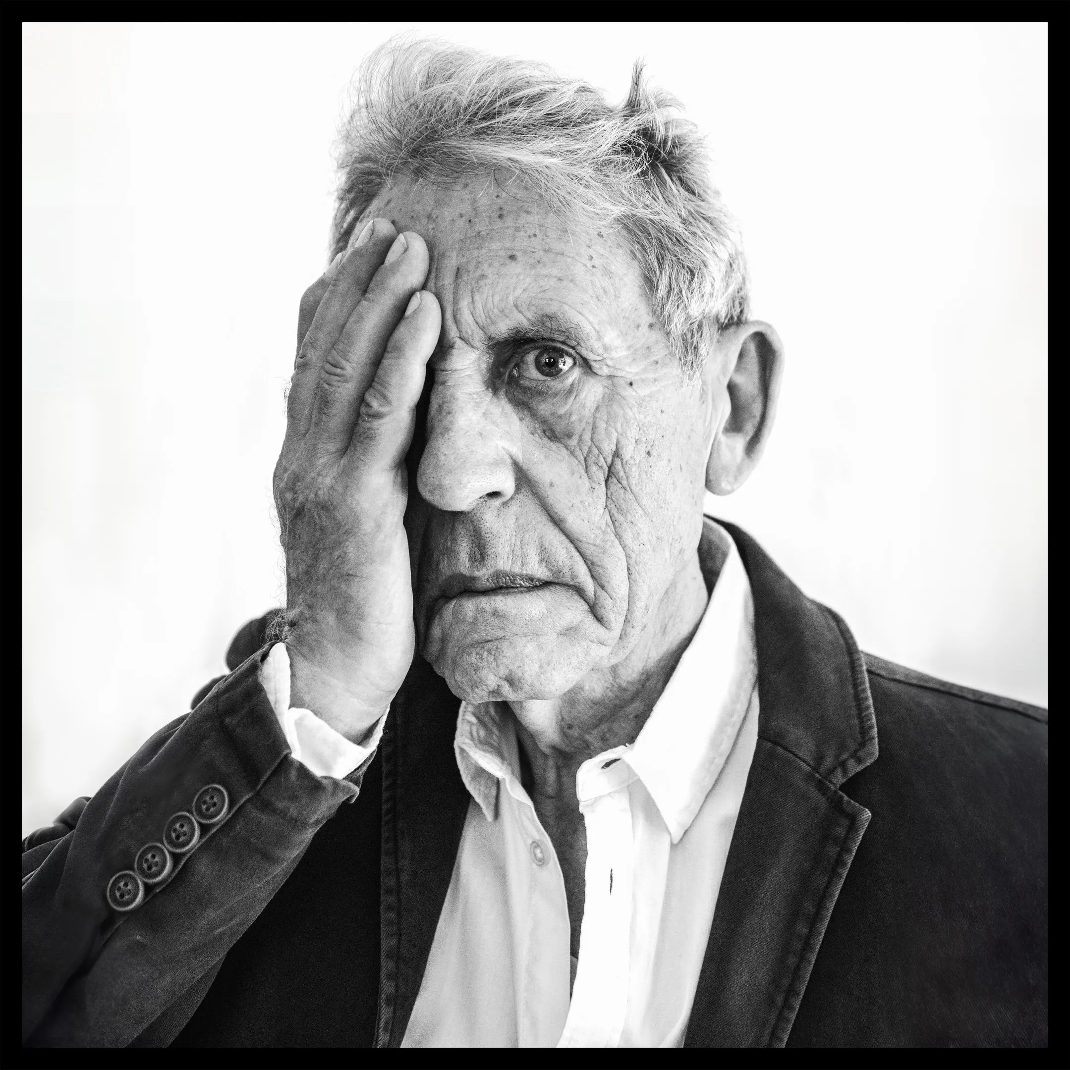 A close portrait of an older man covering one eye with his hand and looking at the camera with the other.