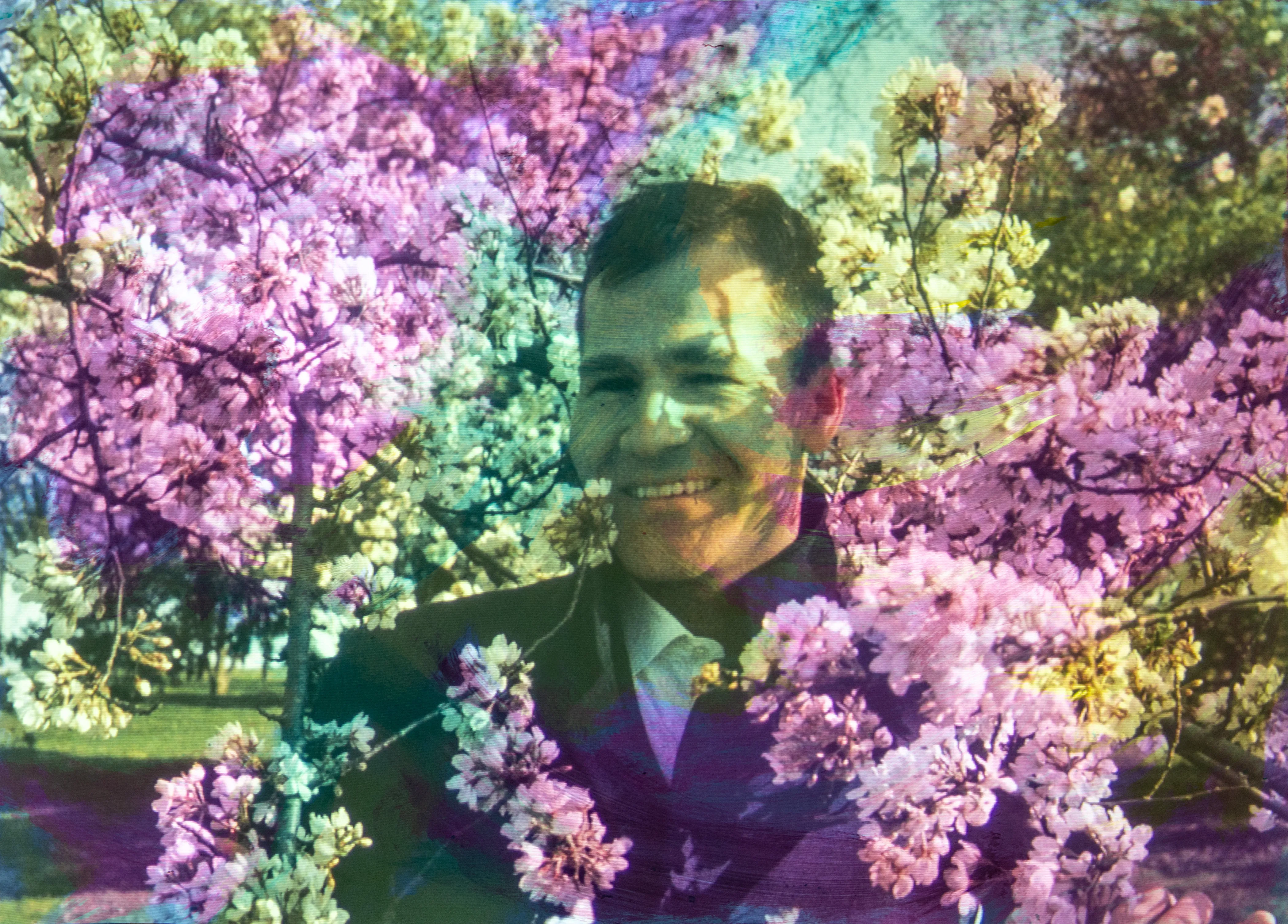 A smiling man in a dark suit stands among pink and purple blossoms.