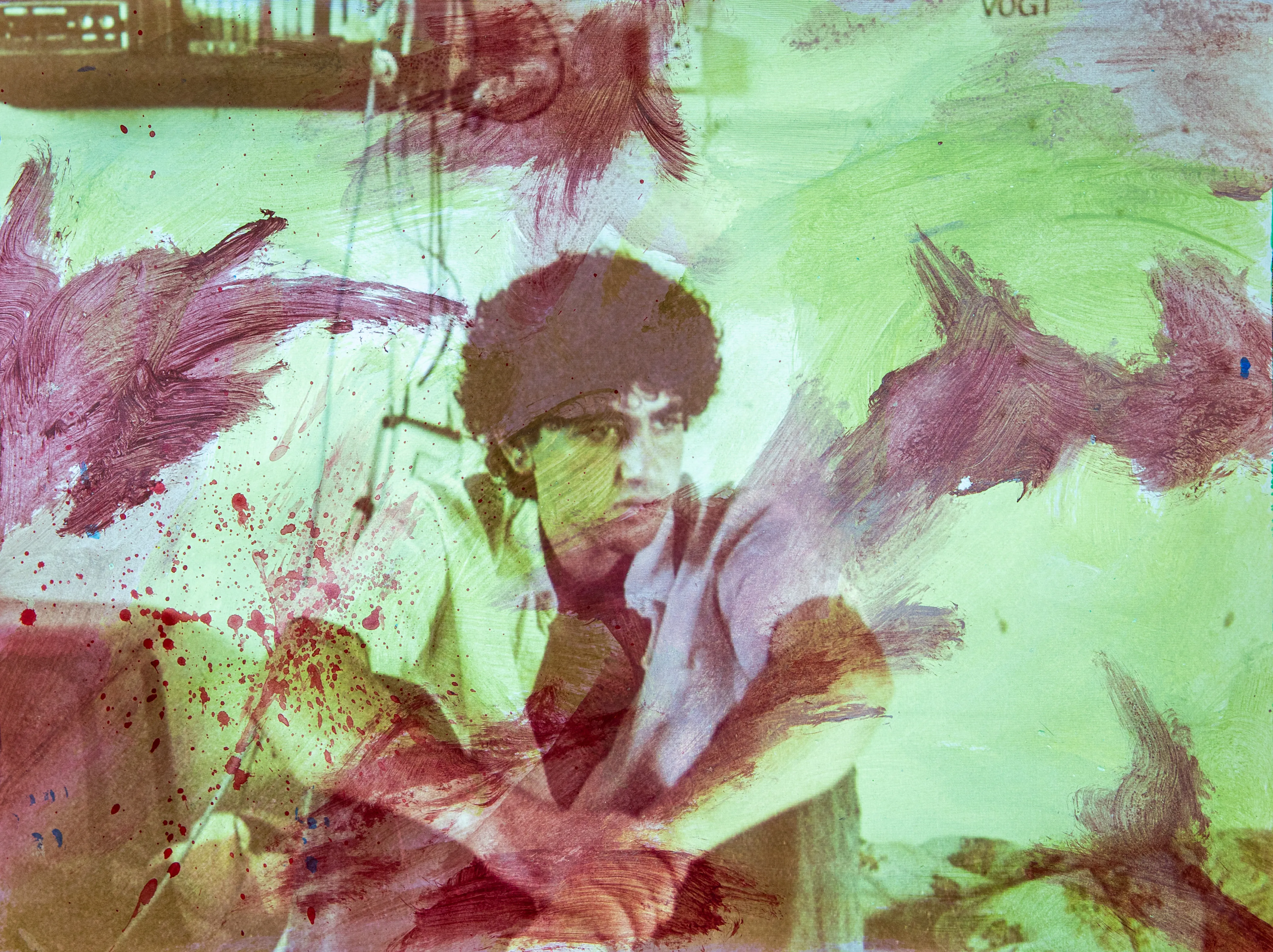 A young man with dark curly hair sits with his arms on his knees against pale green paint and purple strokes.