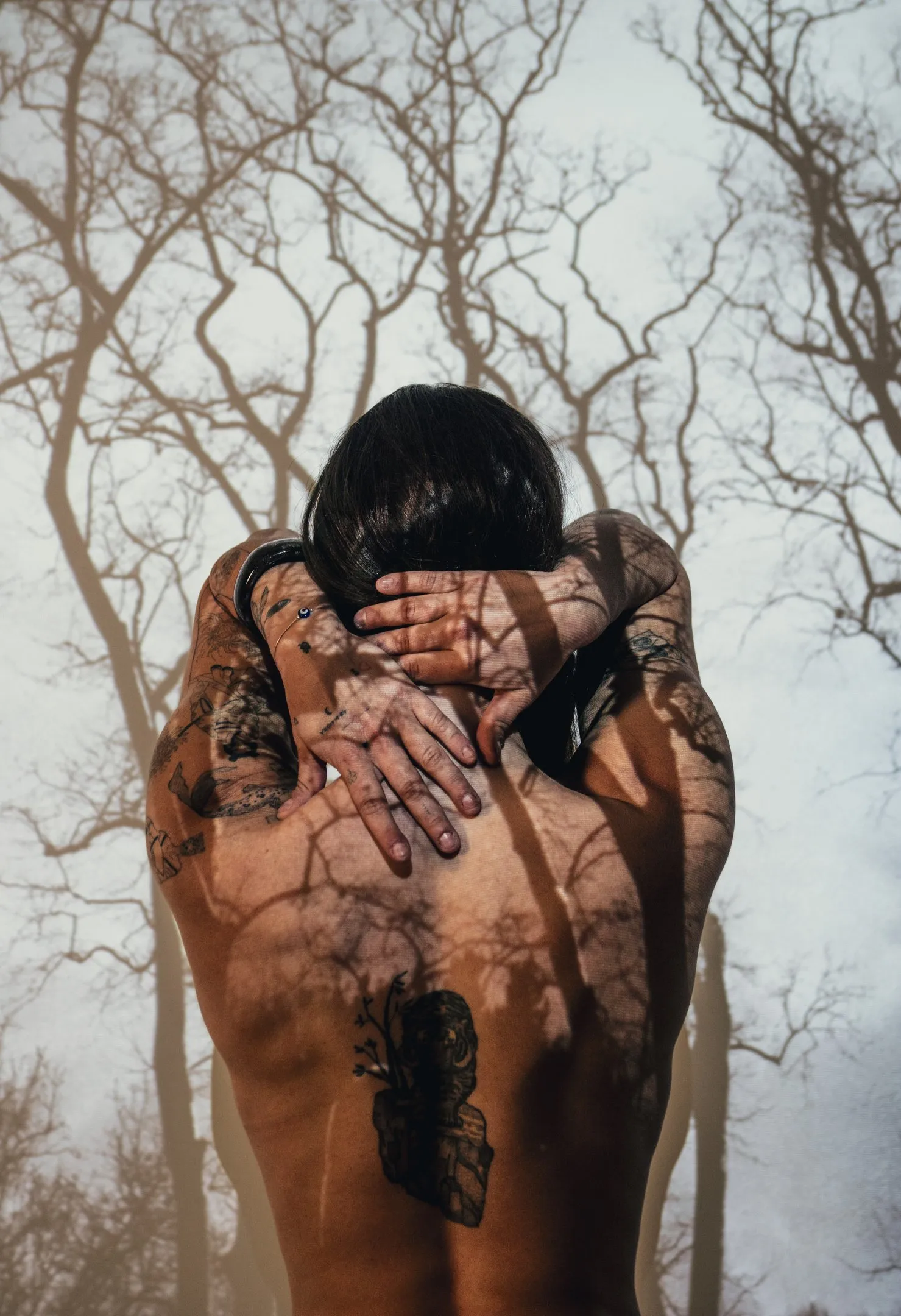 A figure seen from behind with both arms crossed over the shoulders, hands gripping, a tattoo on the back, branch shadows across the skin.
