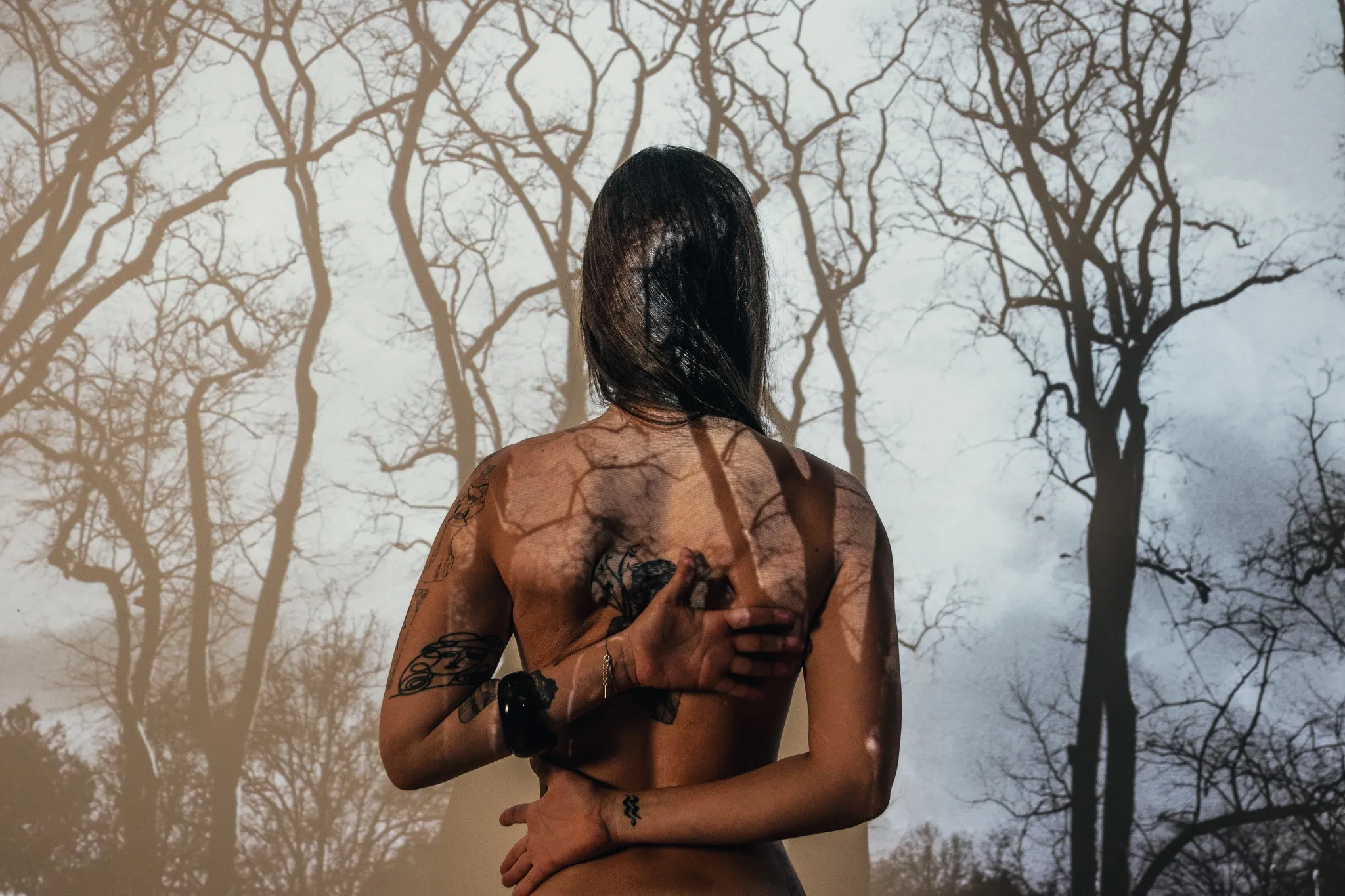 A figure seen from behind with arms bent behind the back, in front of bare trees and a cloudy sky.
