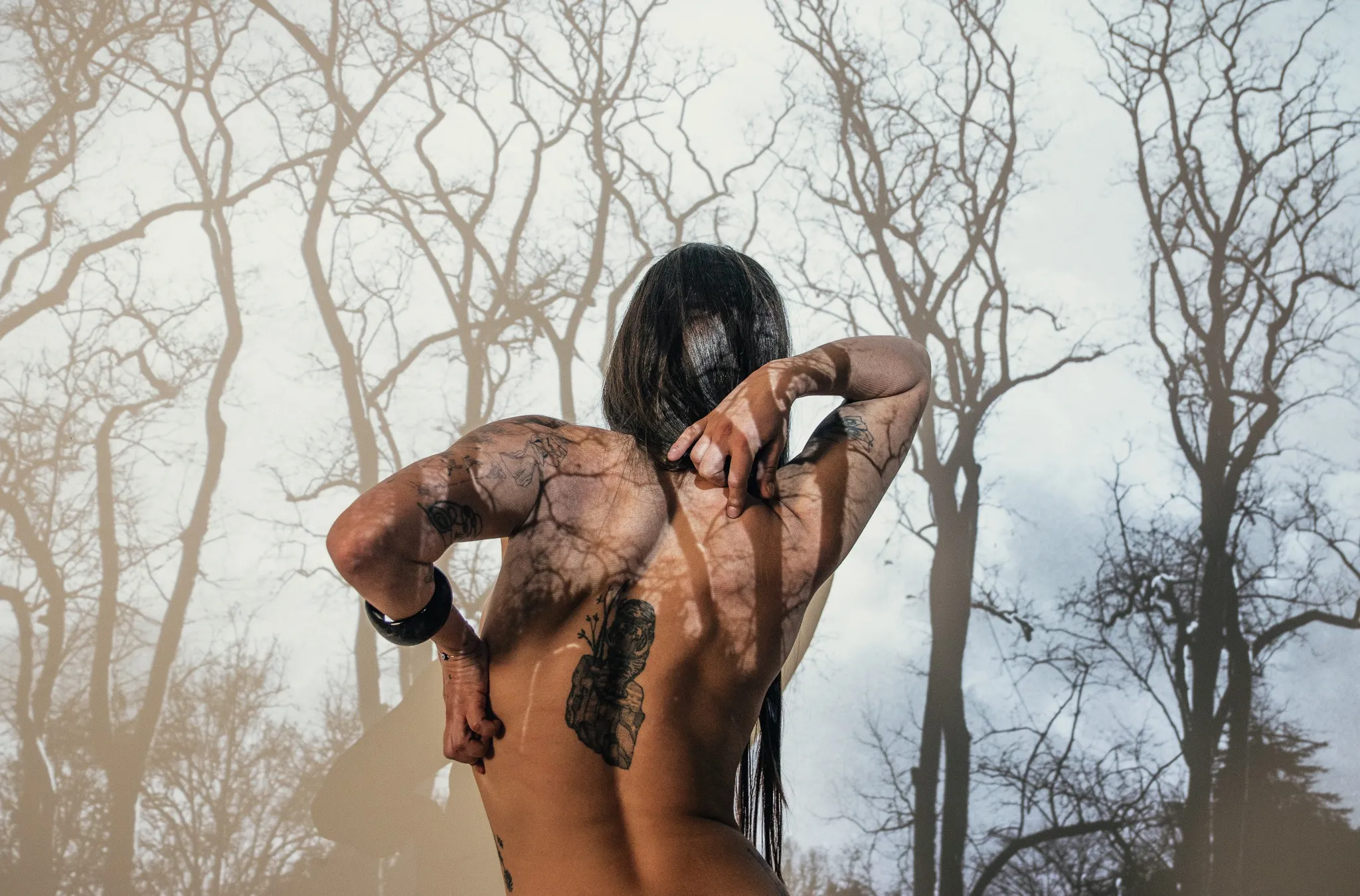 A figure seen from behind with arms raised and hands at the back of the neck, branch shadows across the back.