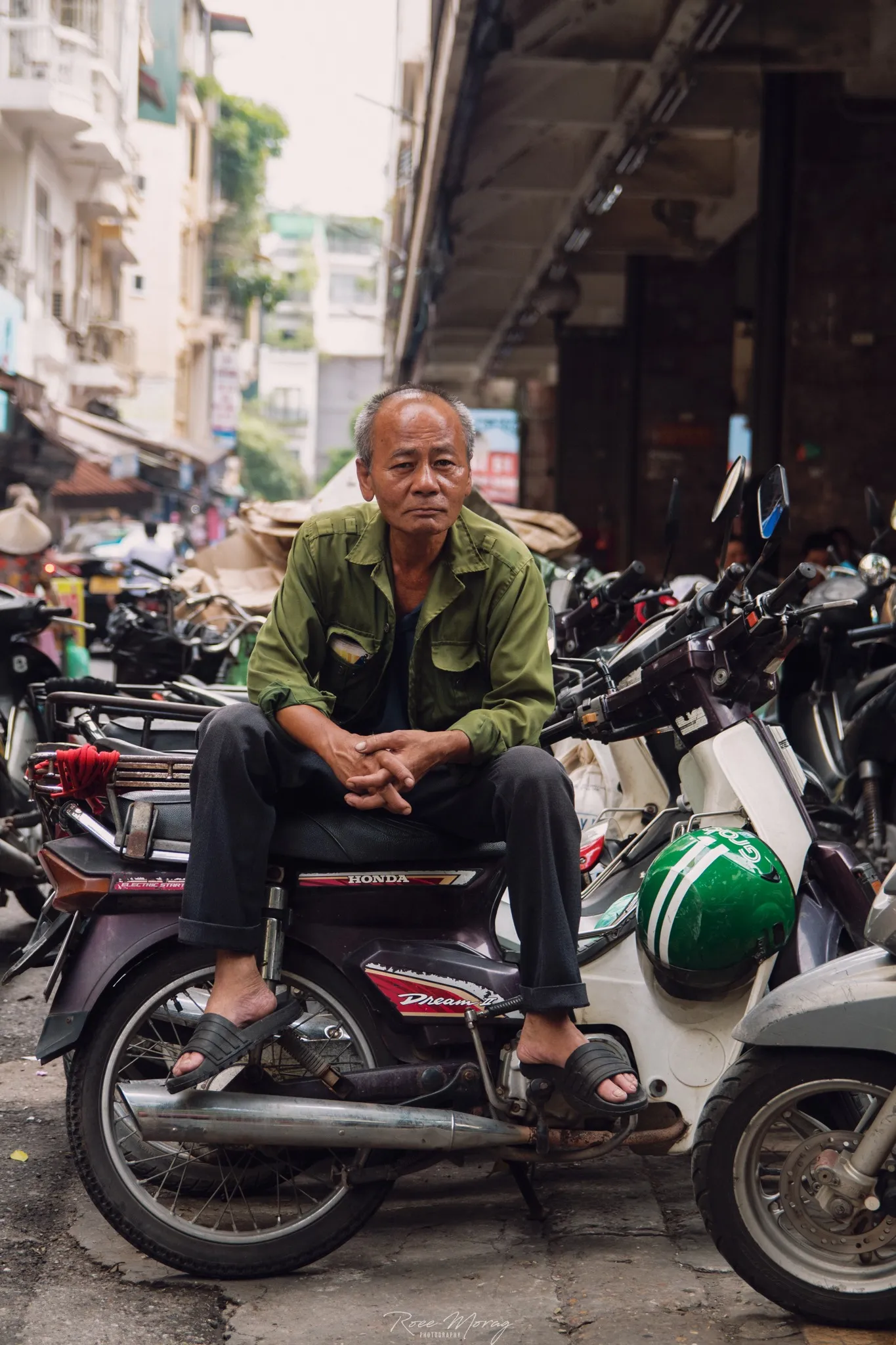 A man in a green jacket sits on a parked motorbike among rows of other motorbikes on a city street.