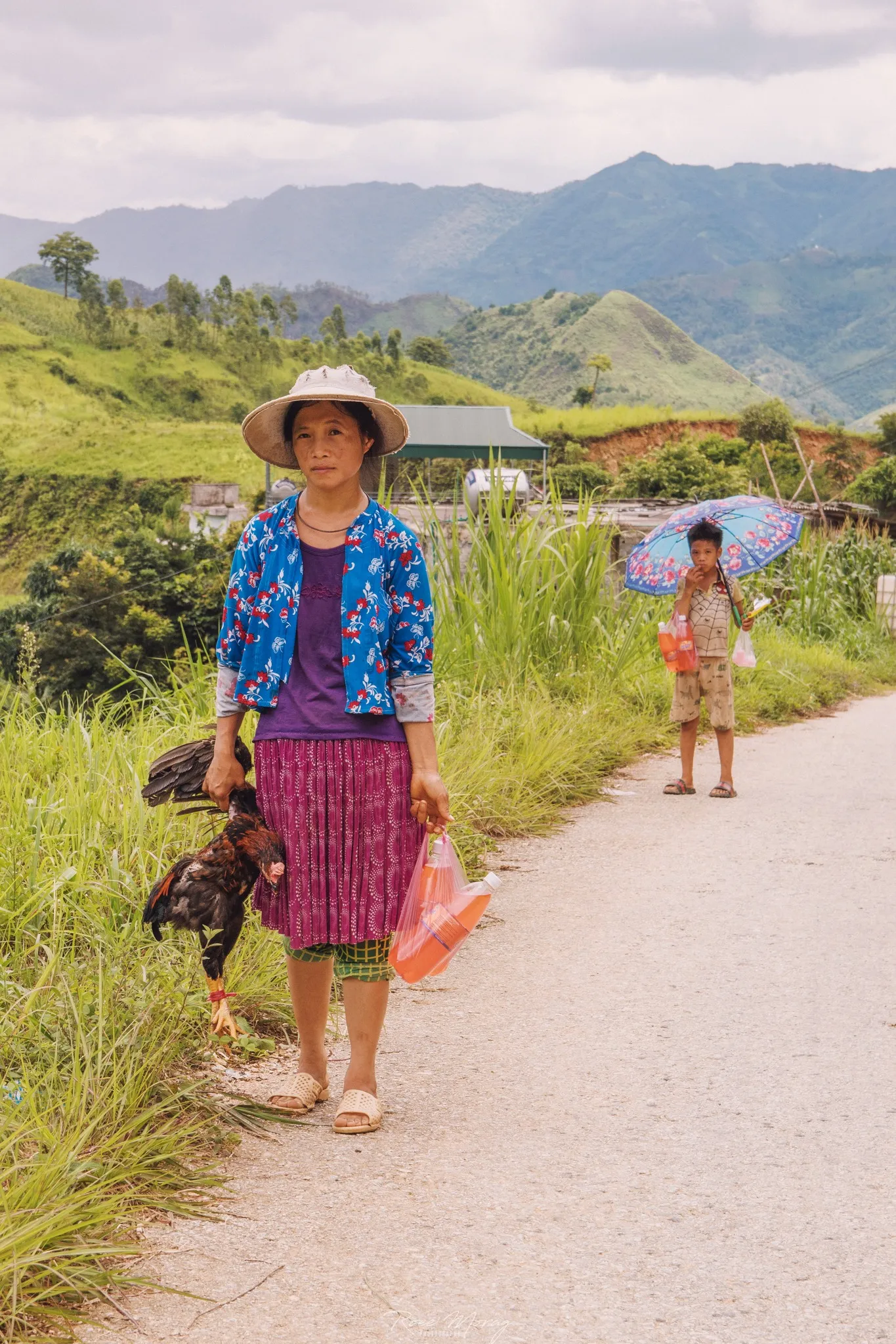 A woman in a wide-brimmed hat and colorful clothes walks along a rural road carrying a chicken, with a child holding an umbrella behind her.