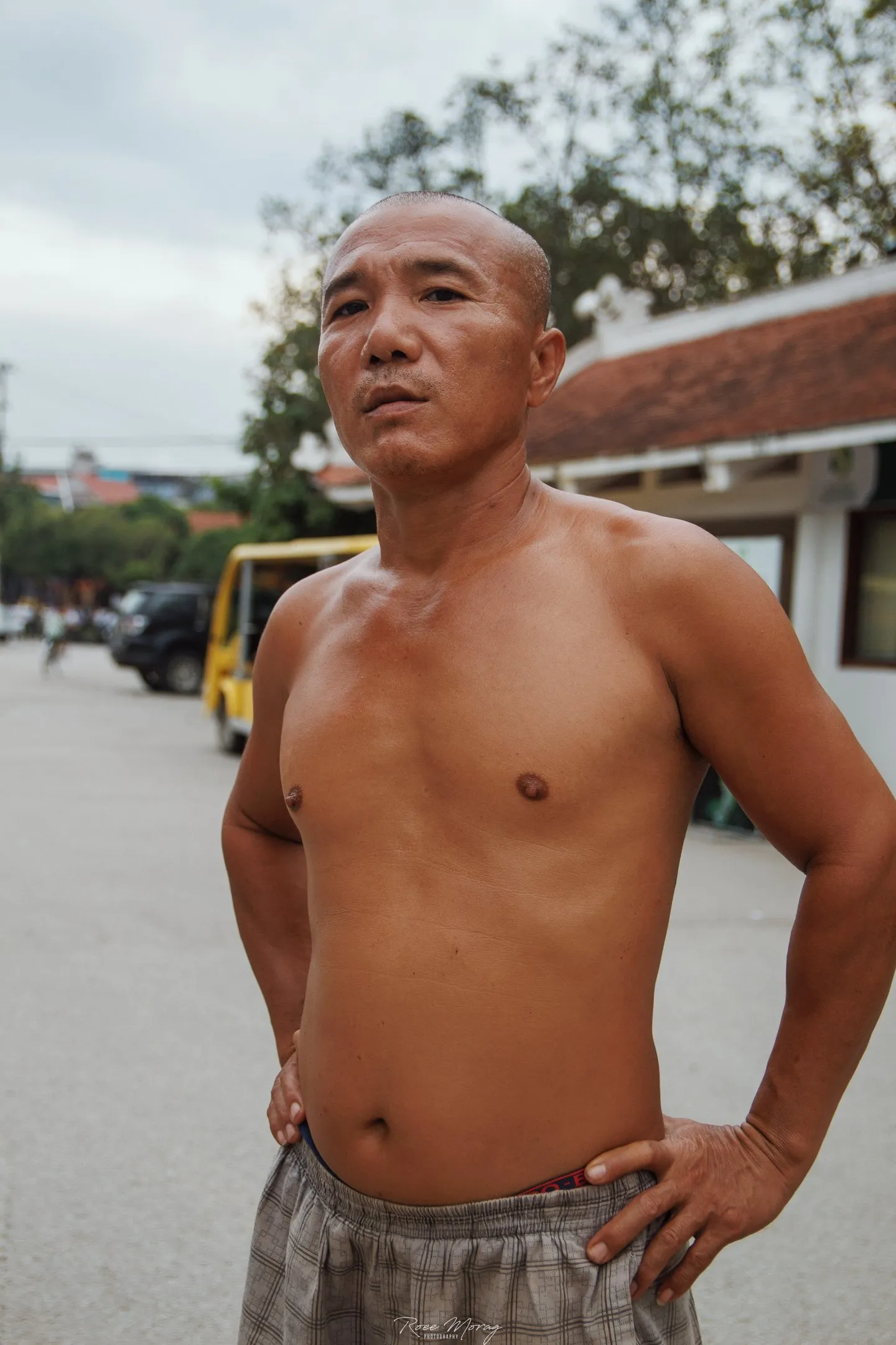 A shirtless man with a shaved head stands with his hands on his hips on a street.