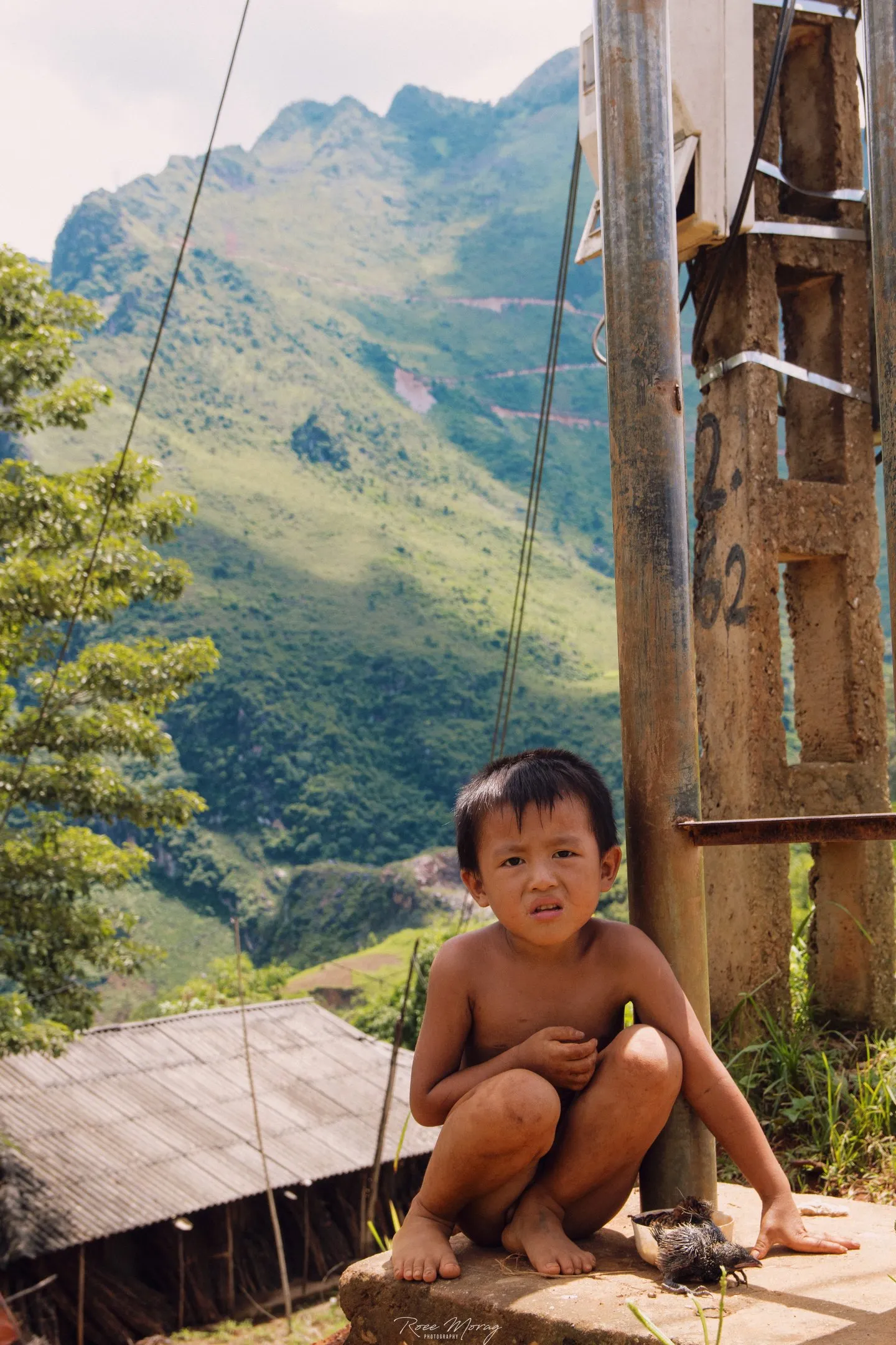 A small child crouches on a stone ledge beside a utility pole, with green mountains behind.