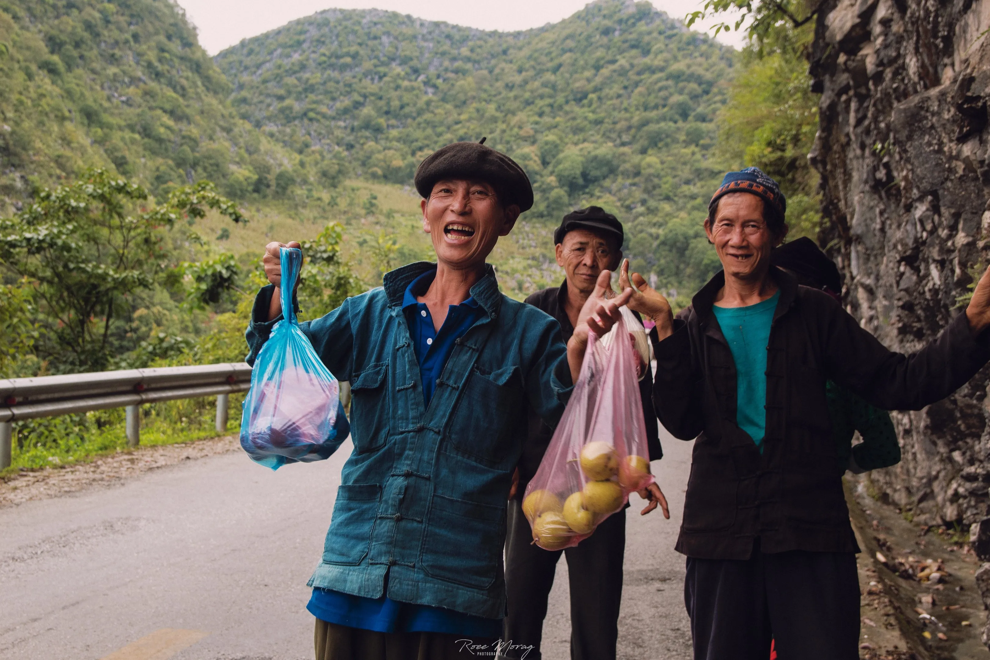 Three smiling men stand on a mountain road, two holding plastic bags of fruit.