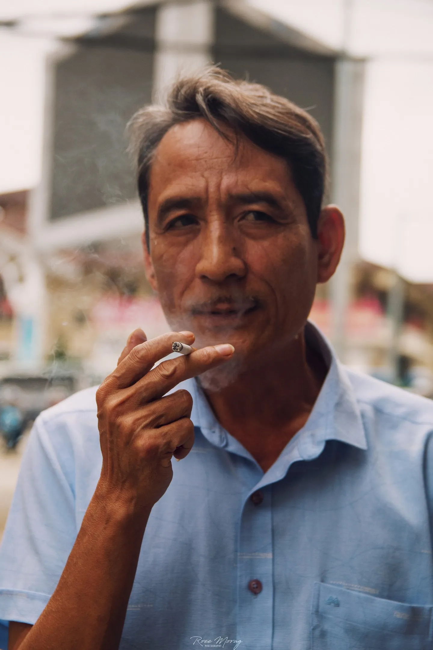 A man in a light blue shirt holds a cigarette to his mouth, with the street blurred behind him.