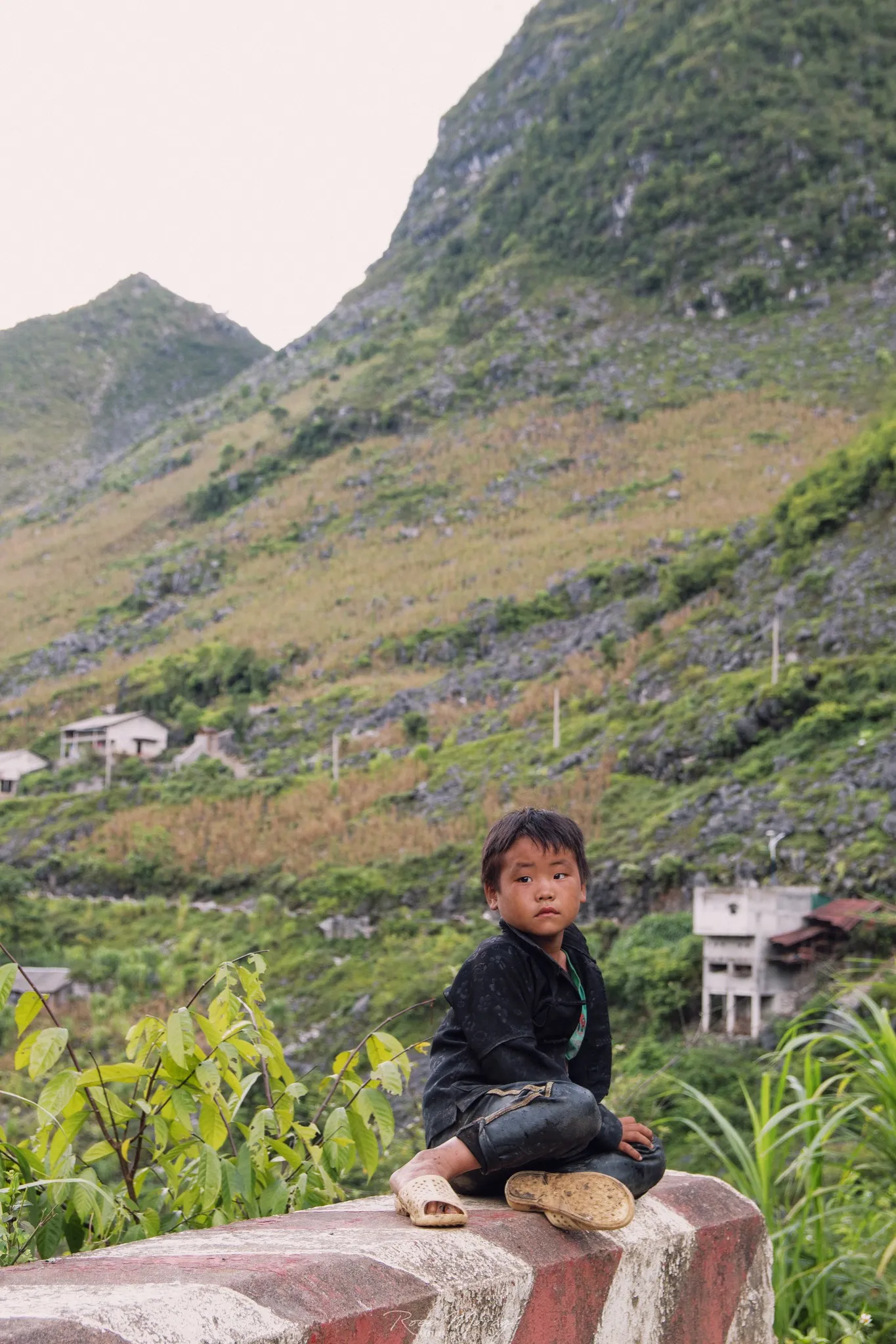 A child sits on a red and white painted roadside barrier, with steep hills behind.