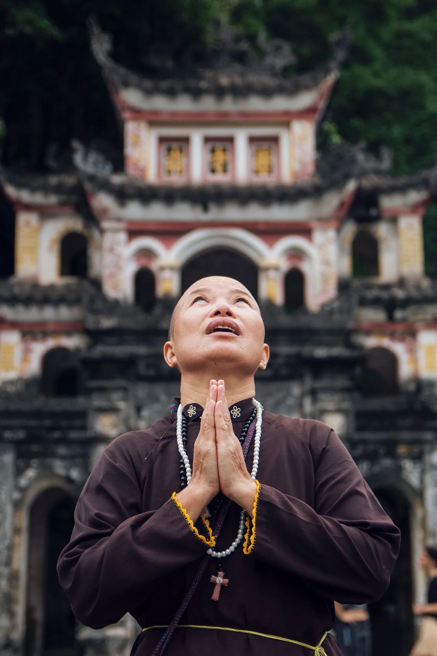 A person in dark robes with hands pressed together looks upward in front of a pagoda-style temple gate.