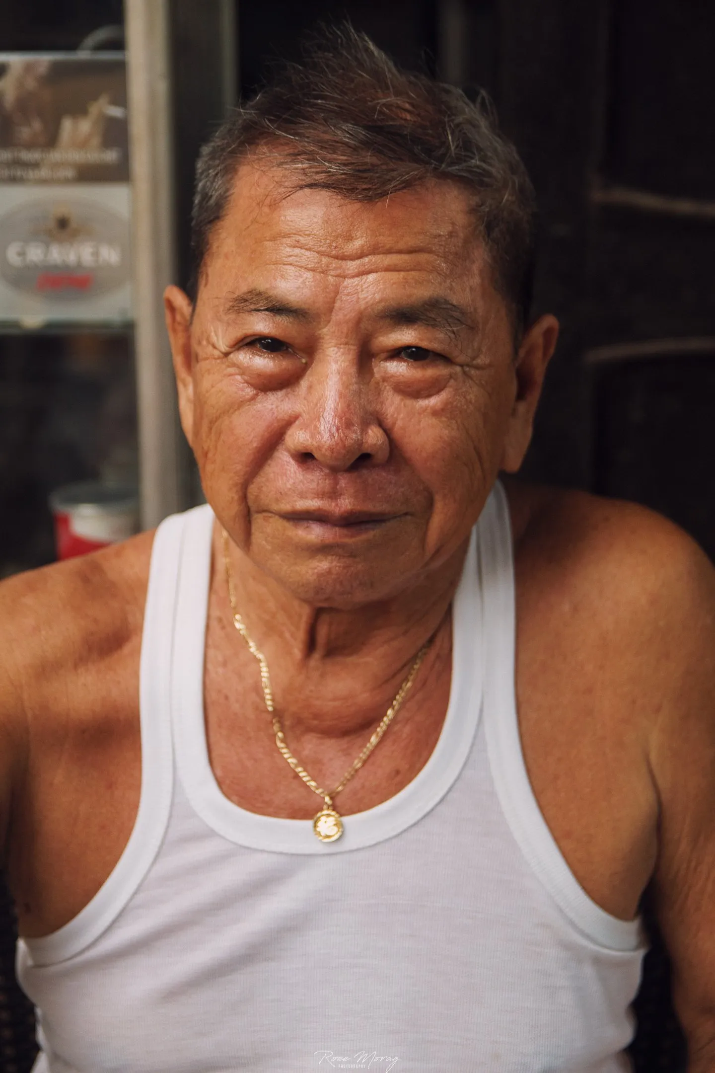 An older man in a white tank top and gold chain looks at the camera in front of a dark interior.