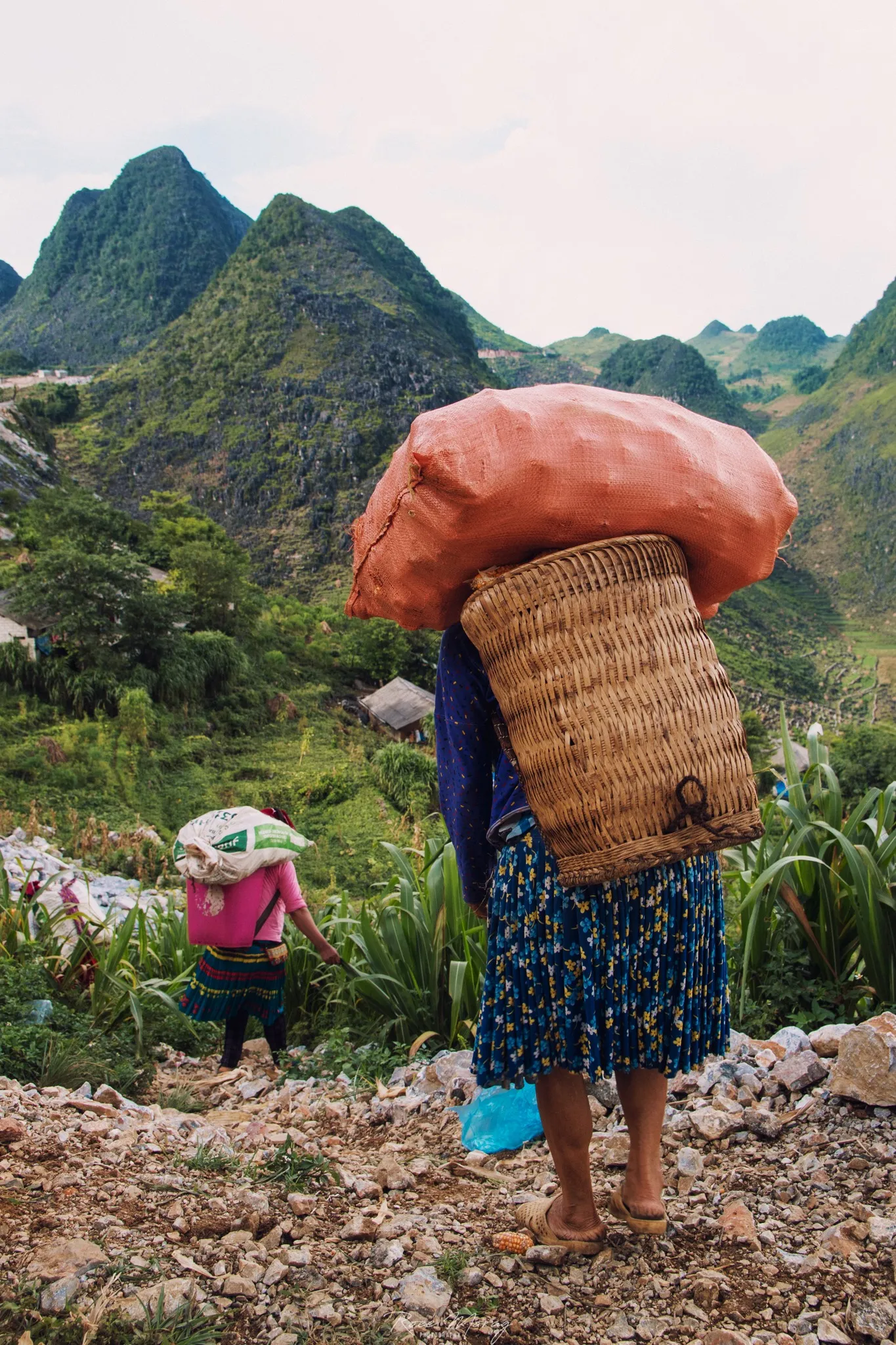 A person in a long patterned skirt carries a woven basket topped with a large orange sack on a rocky path in the hills, with another person in pink carrying a sack in the distance.