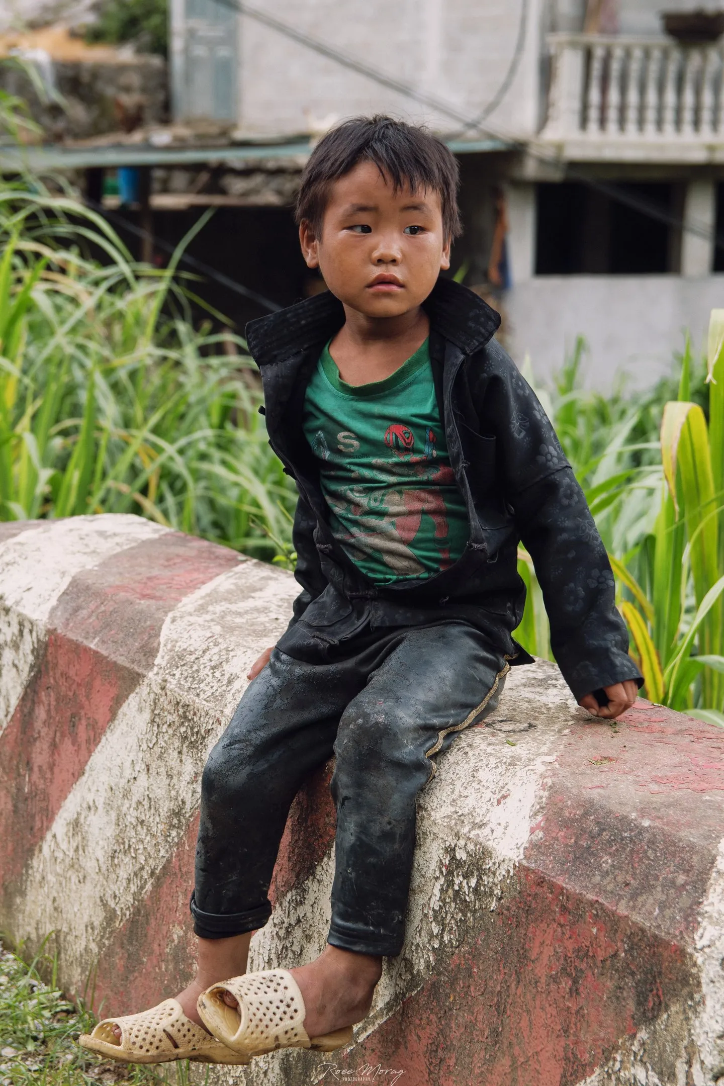 A boy in a dark jacket sits on a red and white painted roadside barrier, with tall grass and a building behind.