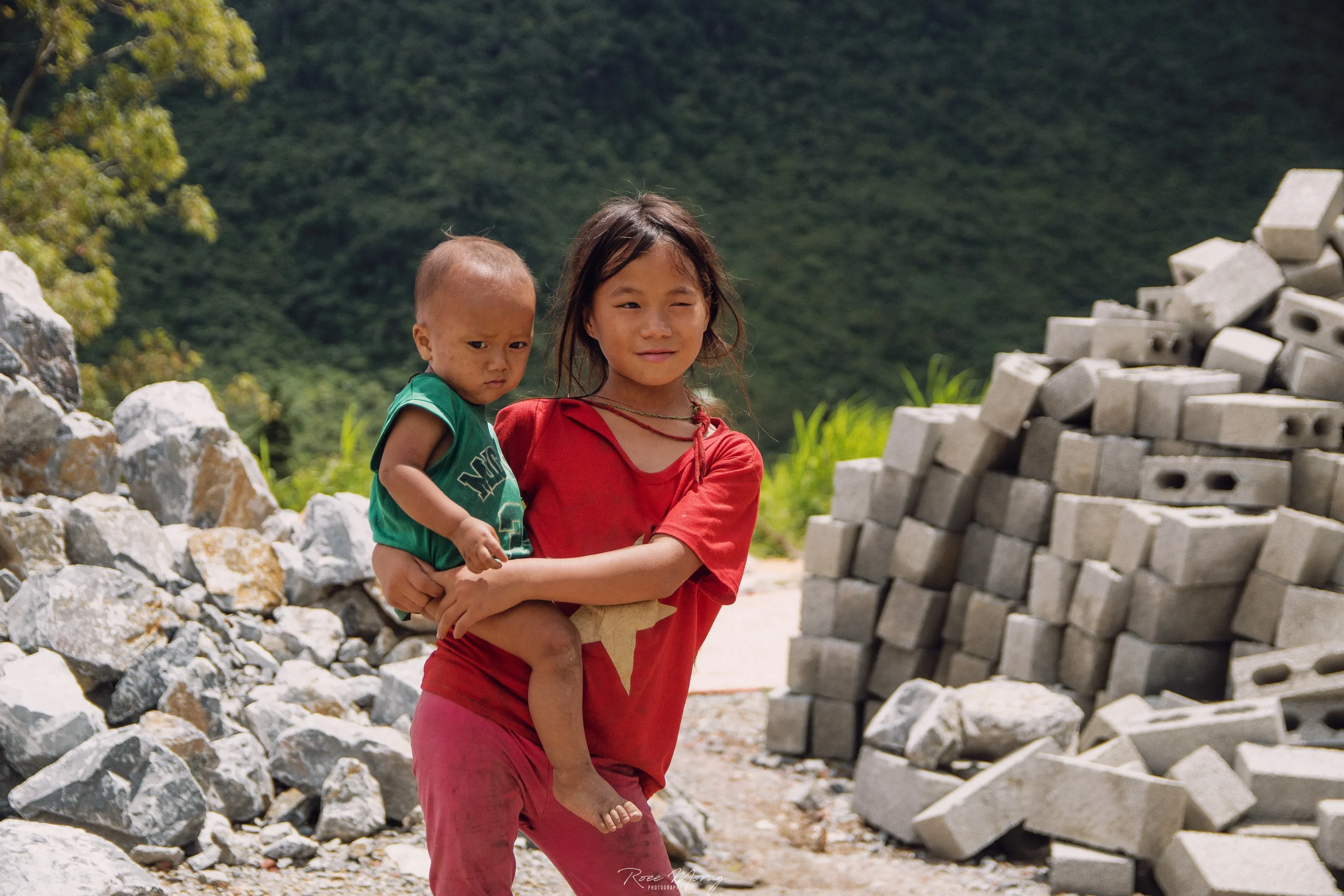 A girl in red holds a toddler in front of piles of rocks and concrete blocks, with forested hills behind.
