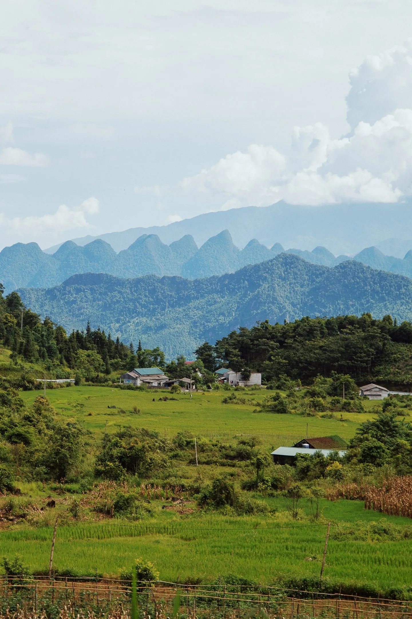 A wide valley of green fields and villages under a cloudy sky, with blue mountains in the distance.