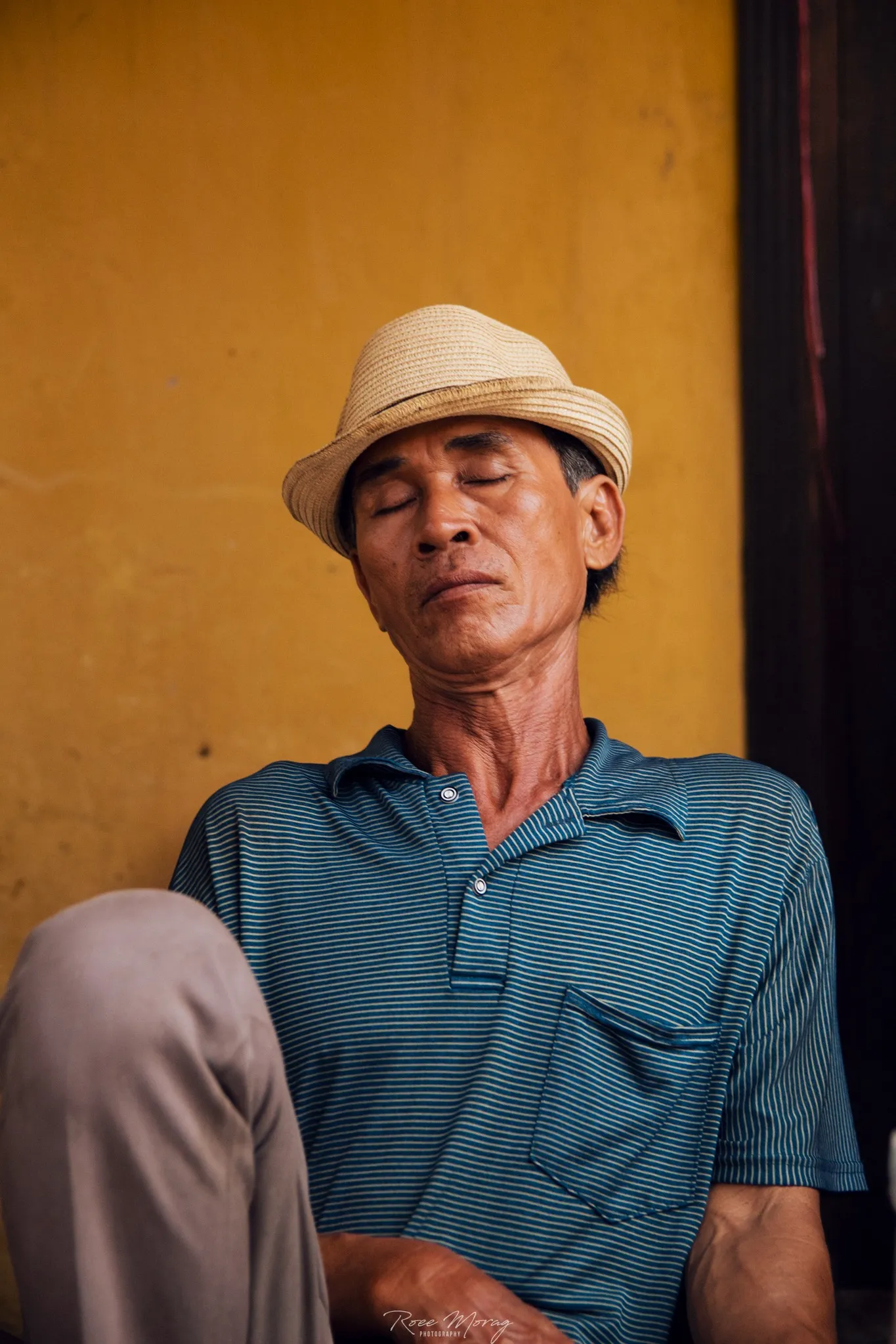 A man in a straw hat and blue polo shirt sits with his eyes closed against a yellow wall.