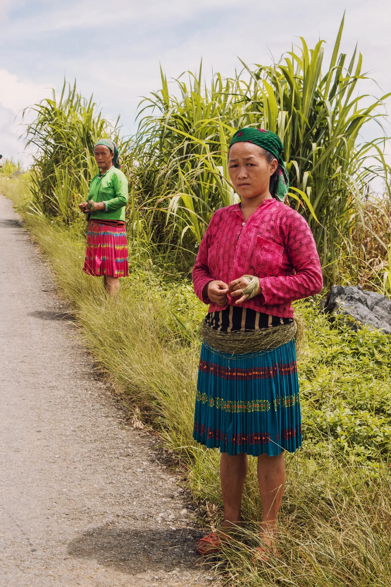 A woman in a pink jacket and bright patterned skirt stands beside a road bordered by tall grass, with another figure further back.
