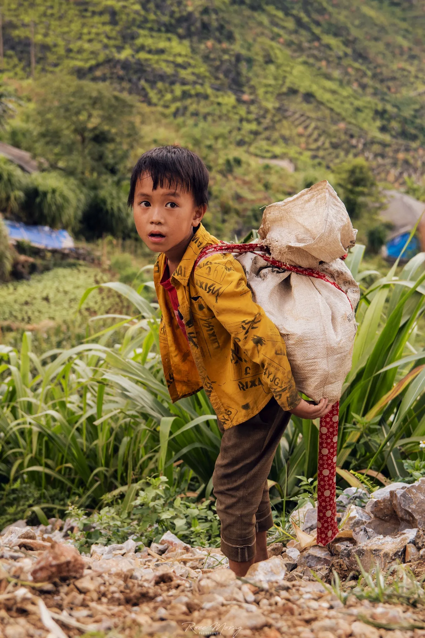 A boy in a yellow jacket carries a sack on his back with a strap across his chest, on a rocky slope, looking back at the camera.